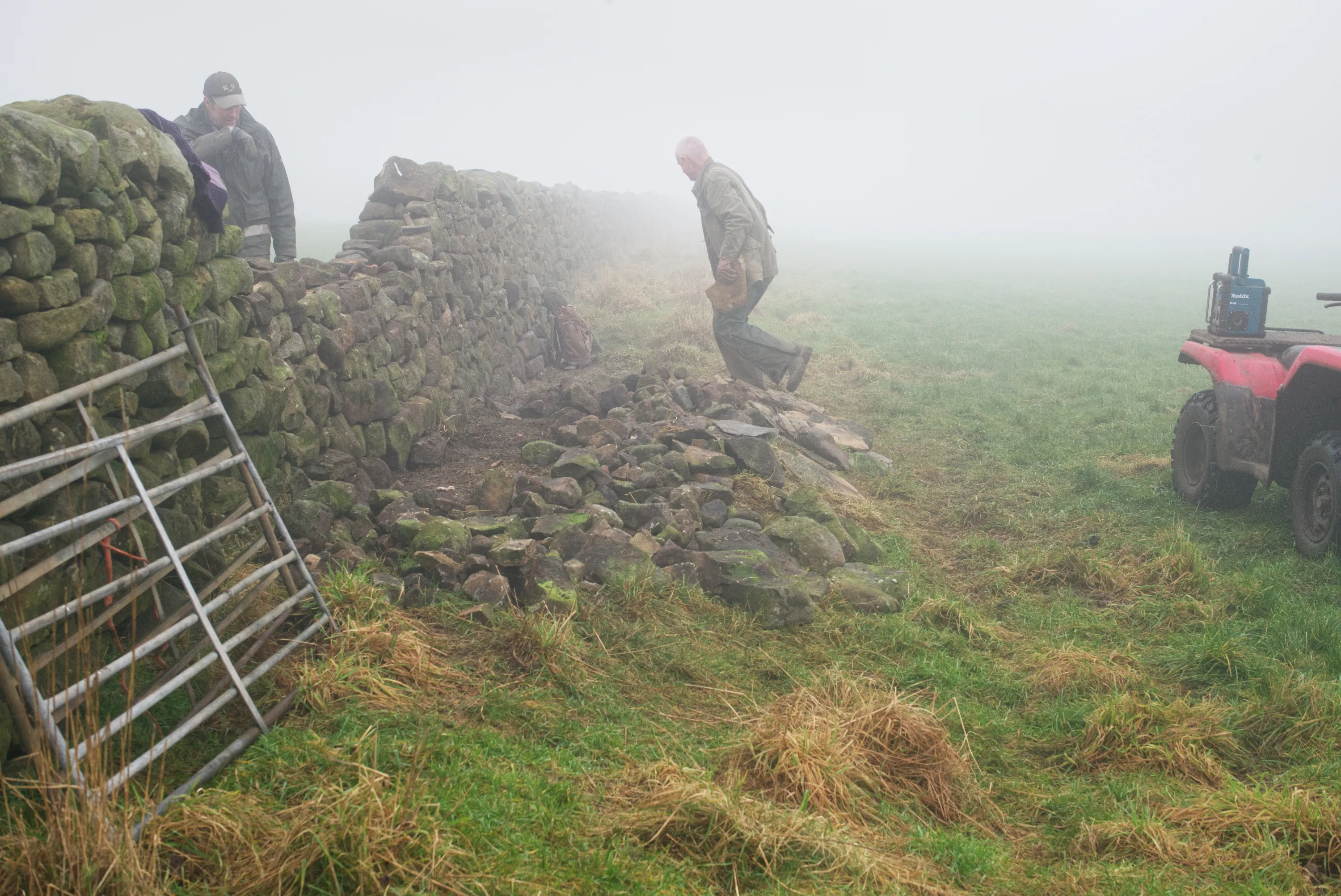Two men next to gap in dry stone wall in fog, one wiping his brow, the other striding