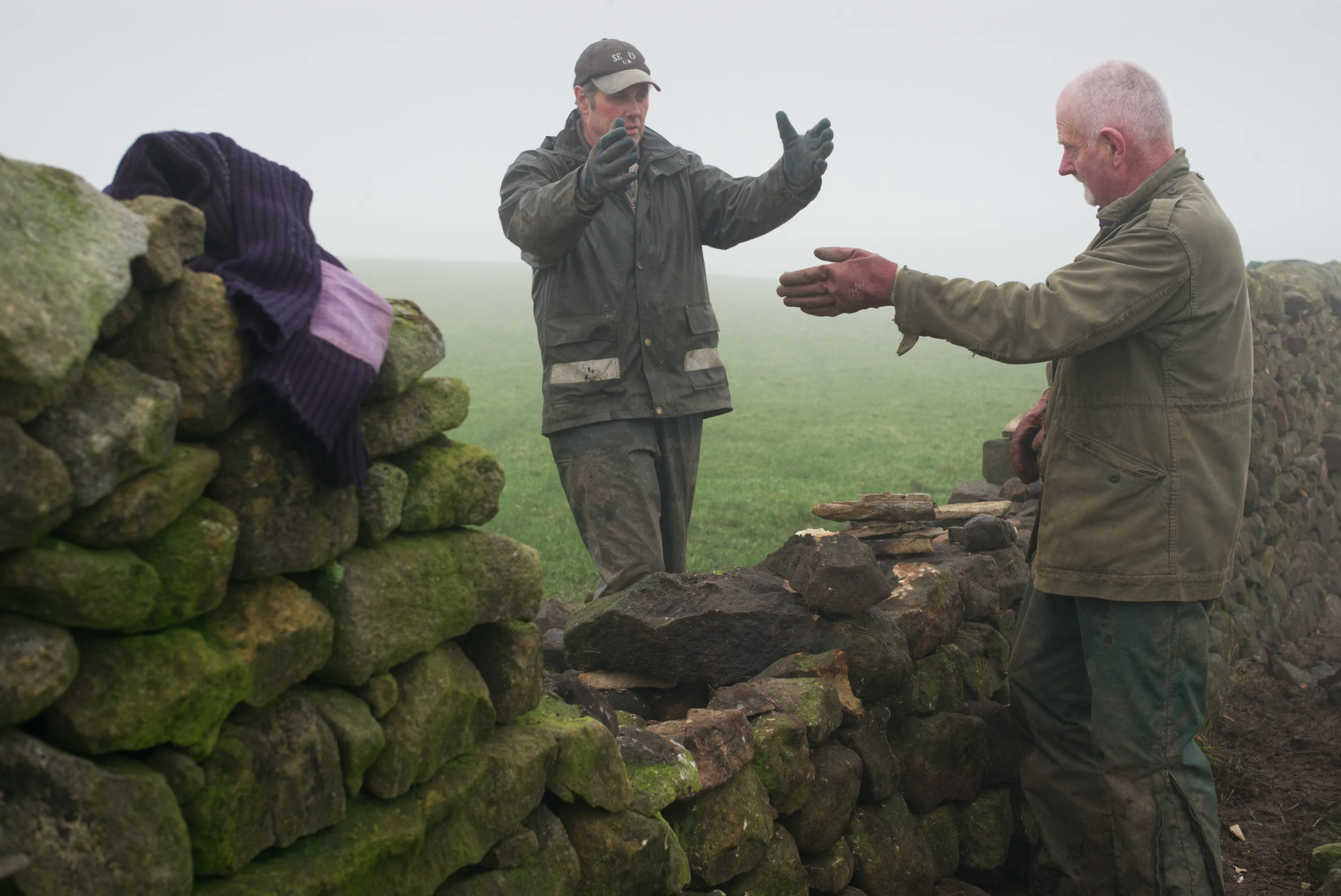 Two men gesturing in conversation beside a dry stone wall in fog