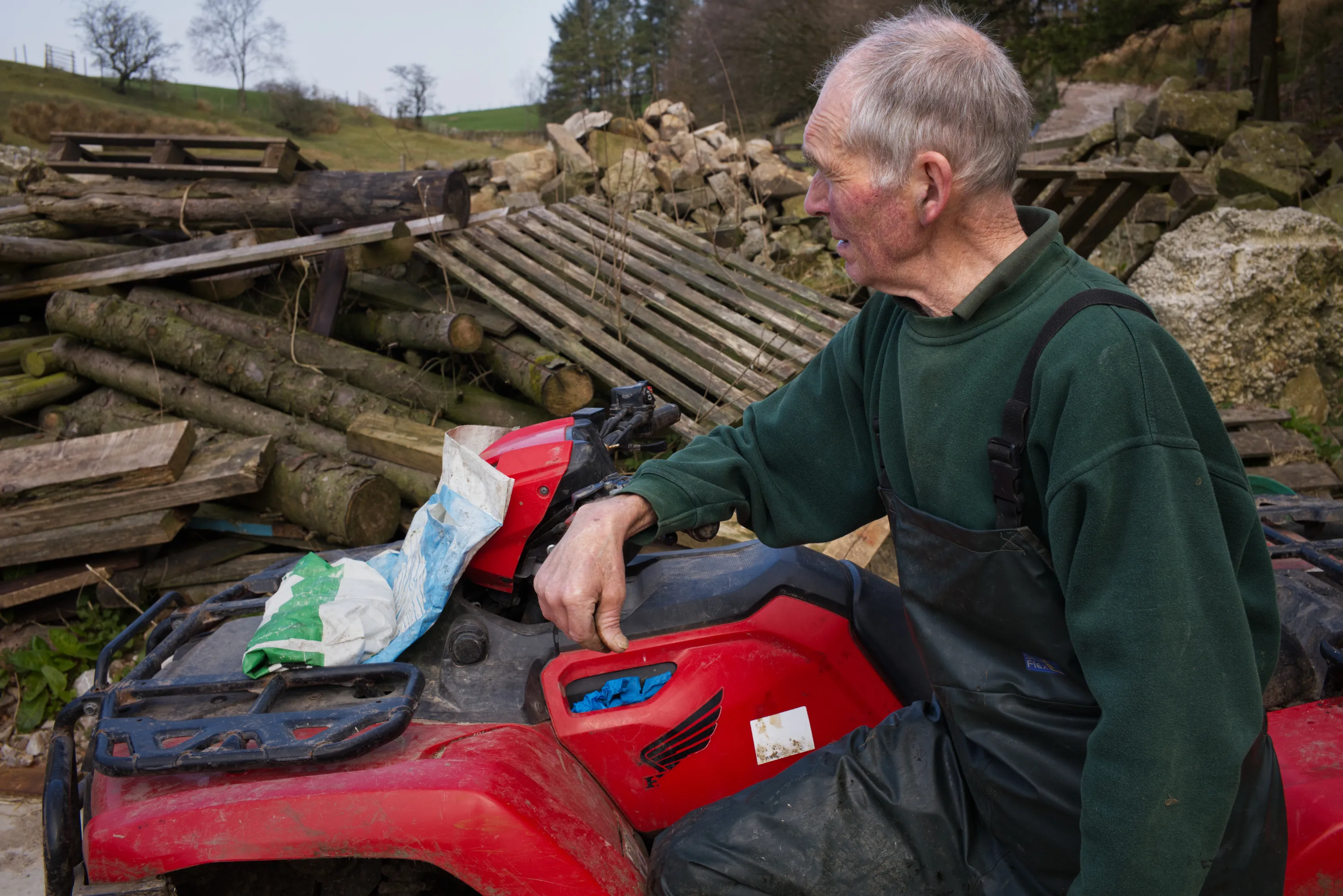 Older man leaning against a red quad bike, farmyard behind him