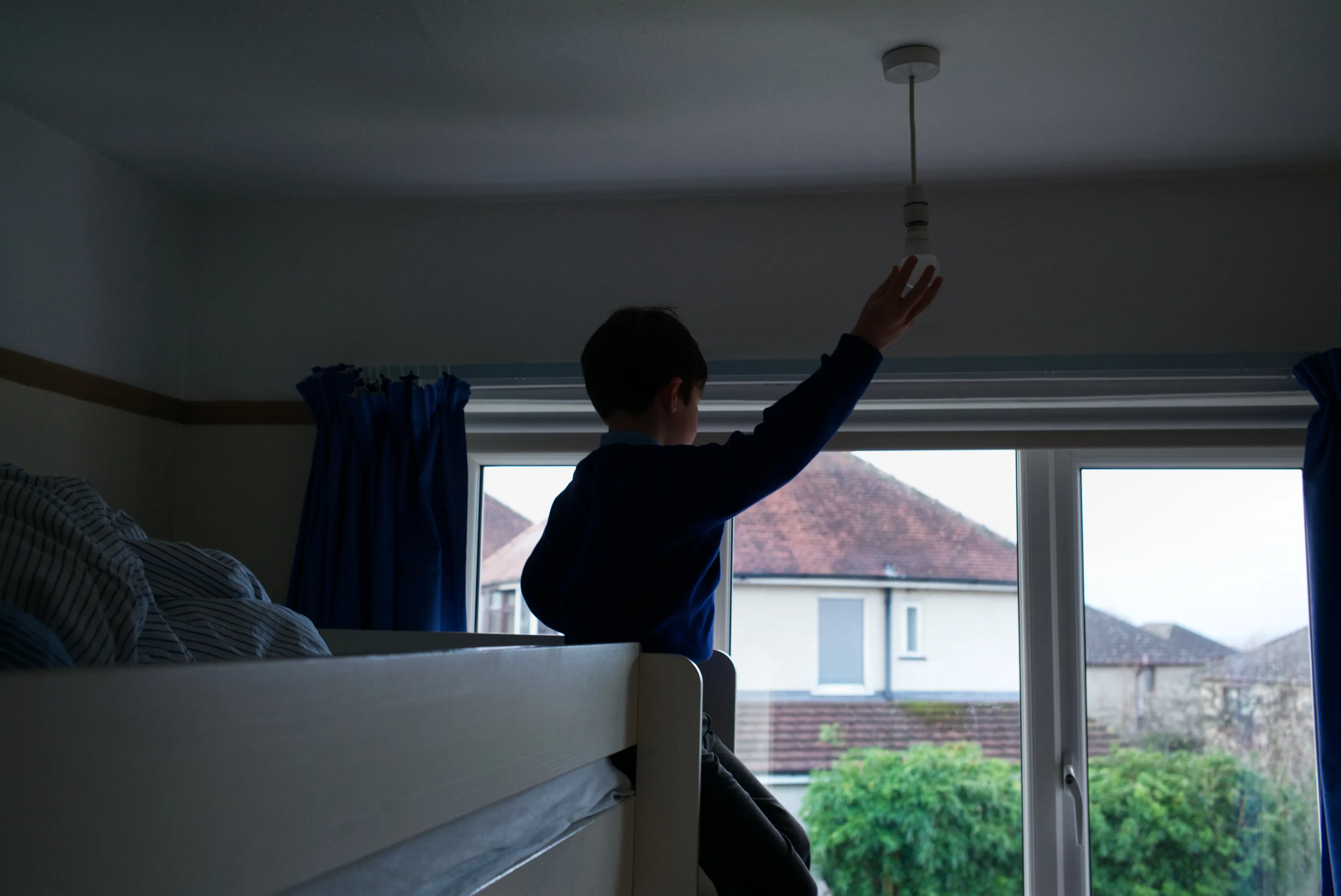 Child silhouetted against a bedroom window, arm raised toward a pendant light fitting, bunk bed beside them