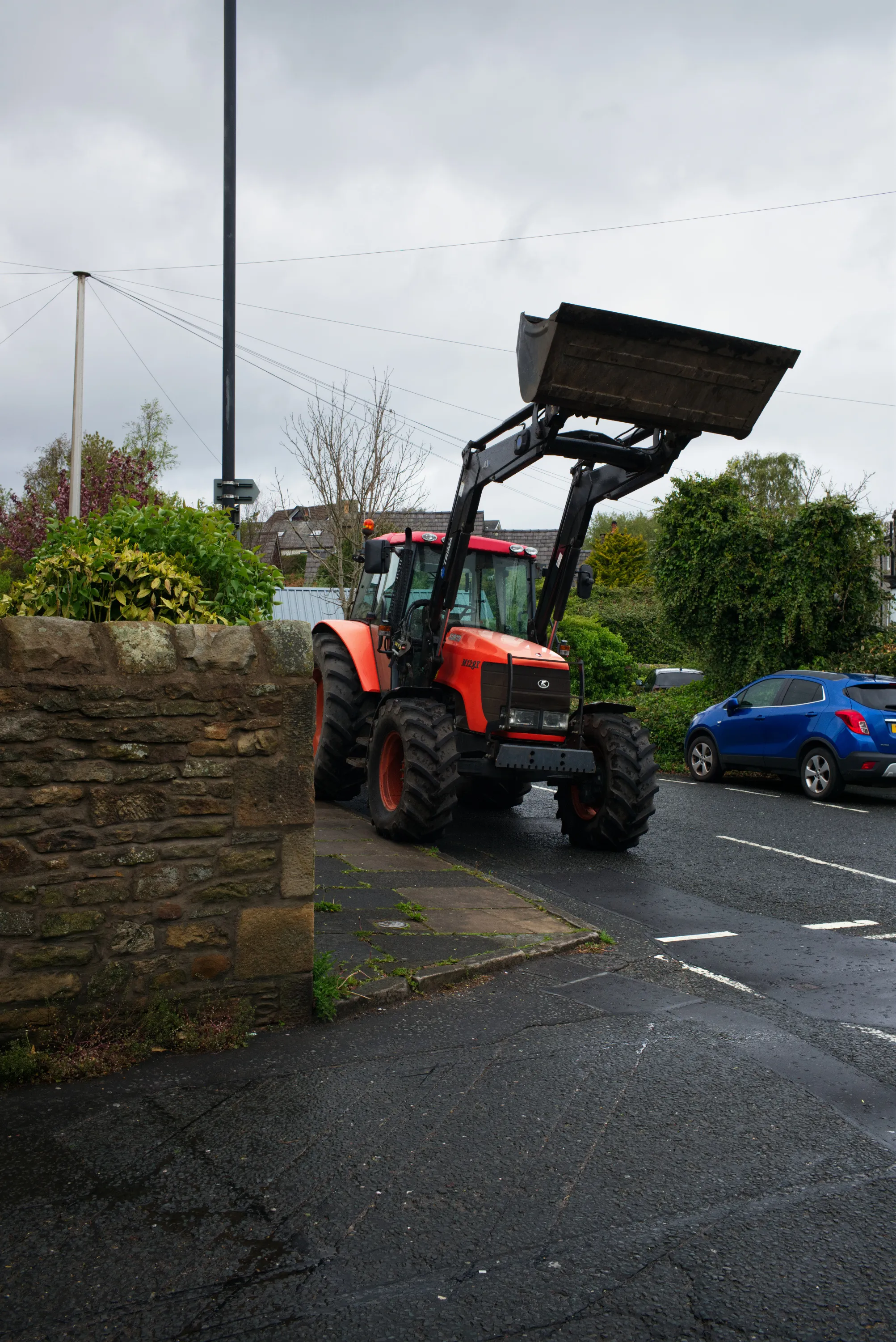 Red front-loader tractor parked on a wet residential street beside a low stone wall