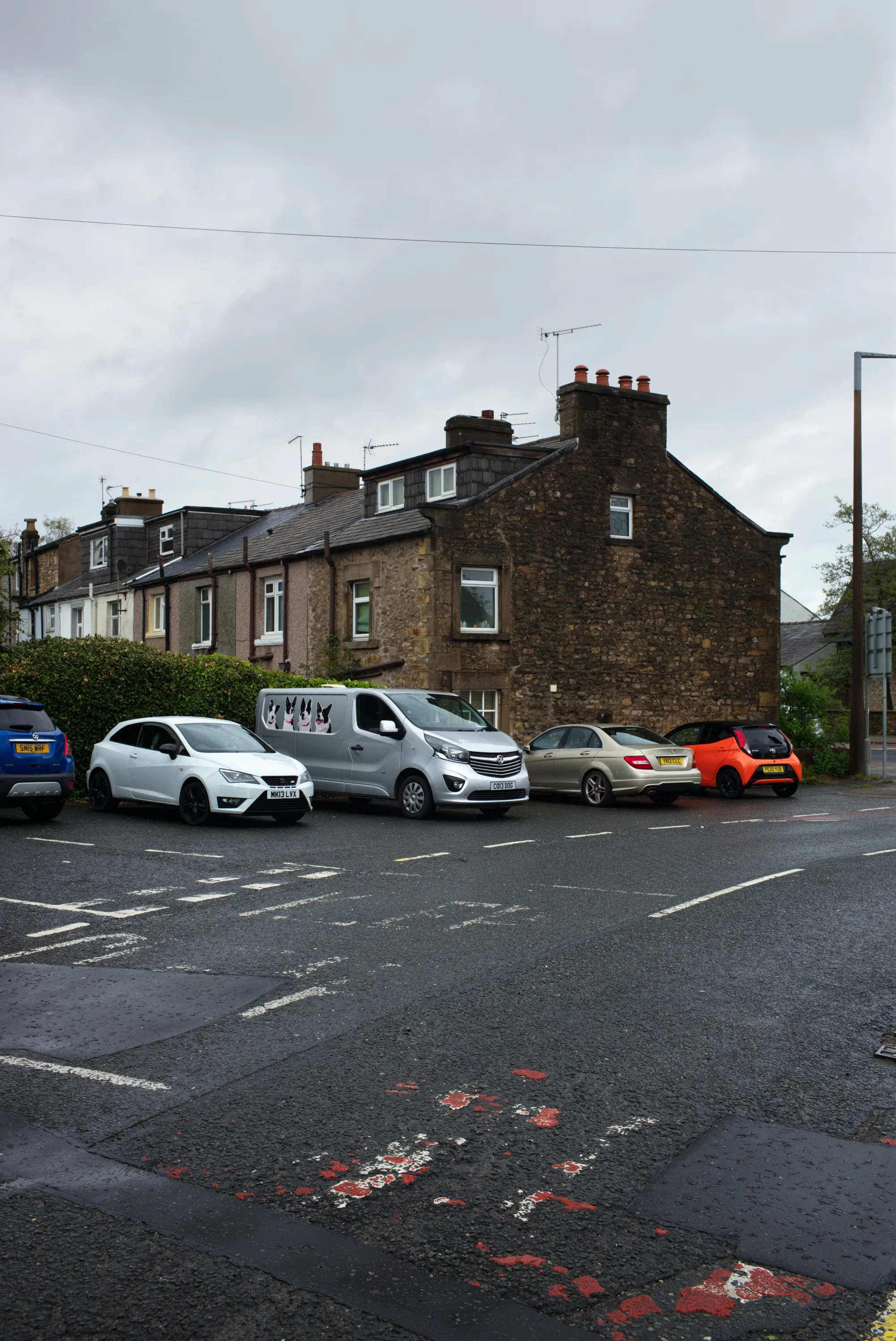Row of parked cars on a wet street in front of stone terraced houses under overcast sky