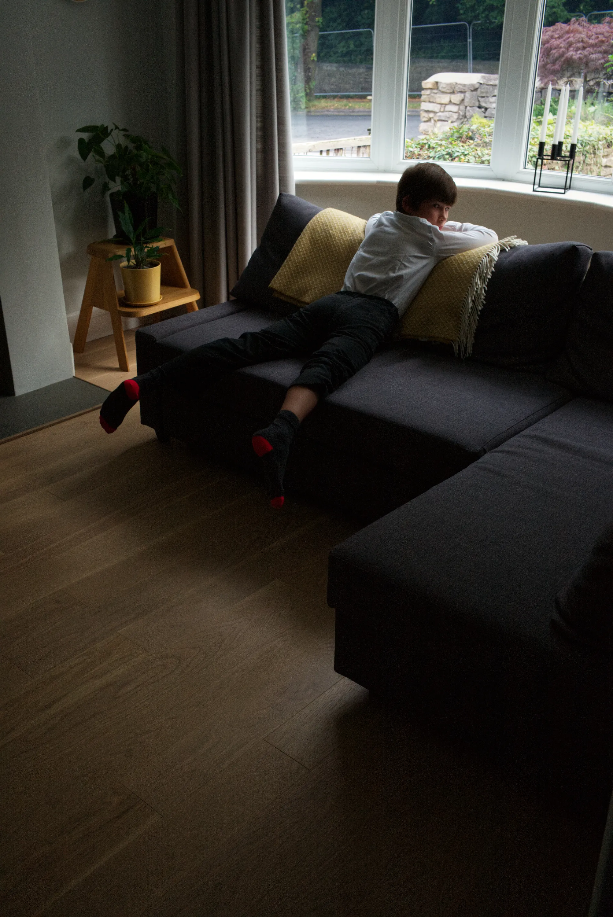 Child lying face-down on a dark sofa, red-heeled socks visible, window with garden view in background