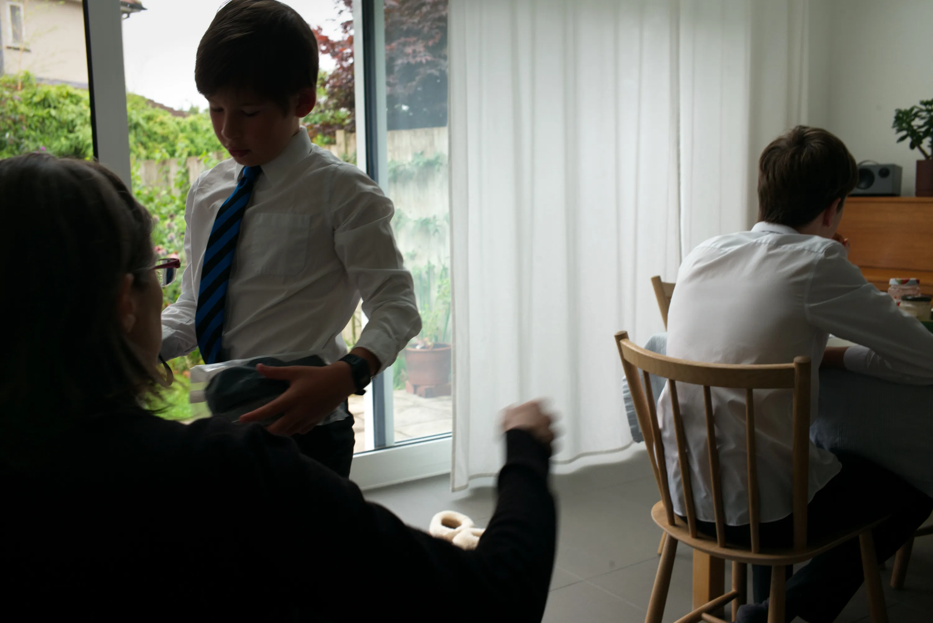 Boy in school uniform handing something to a woman, another child seated at a table near glass doors