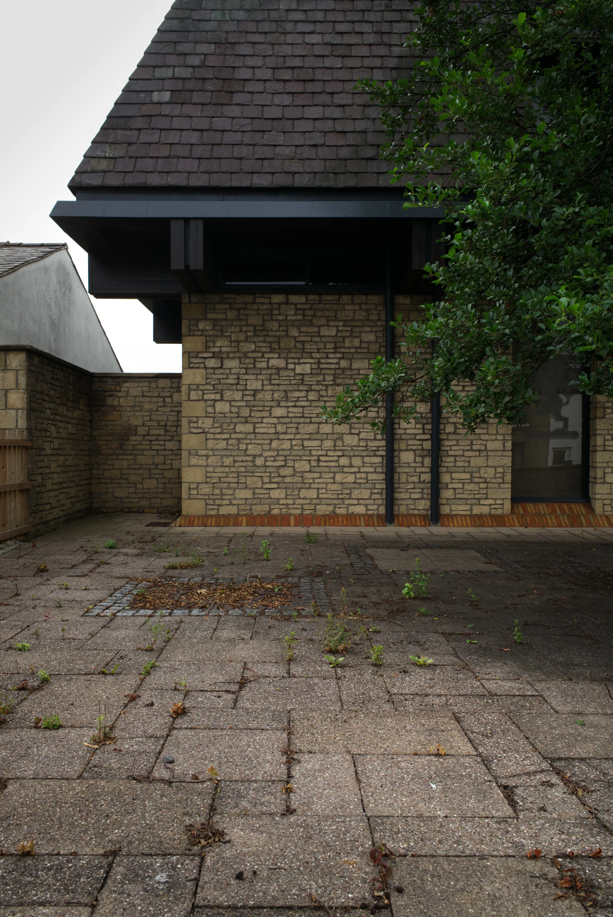 Empty paved courtyard in front of a brick building with a dark cantilevered roof and a tree