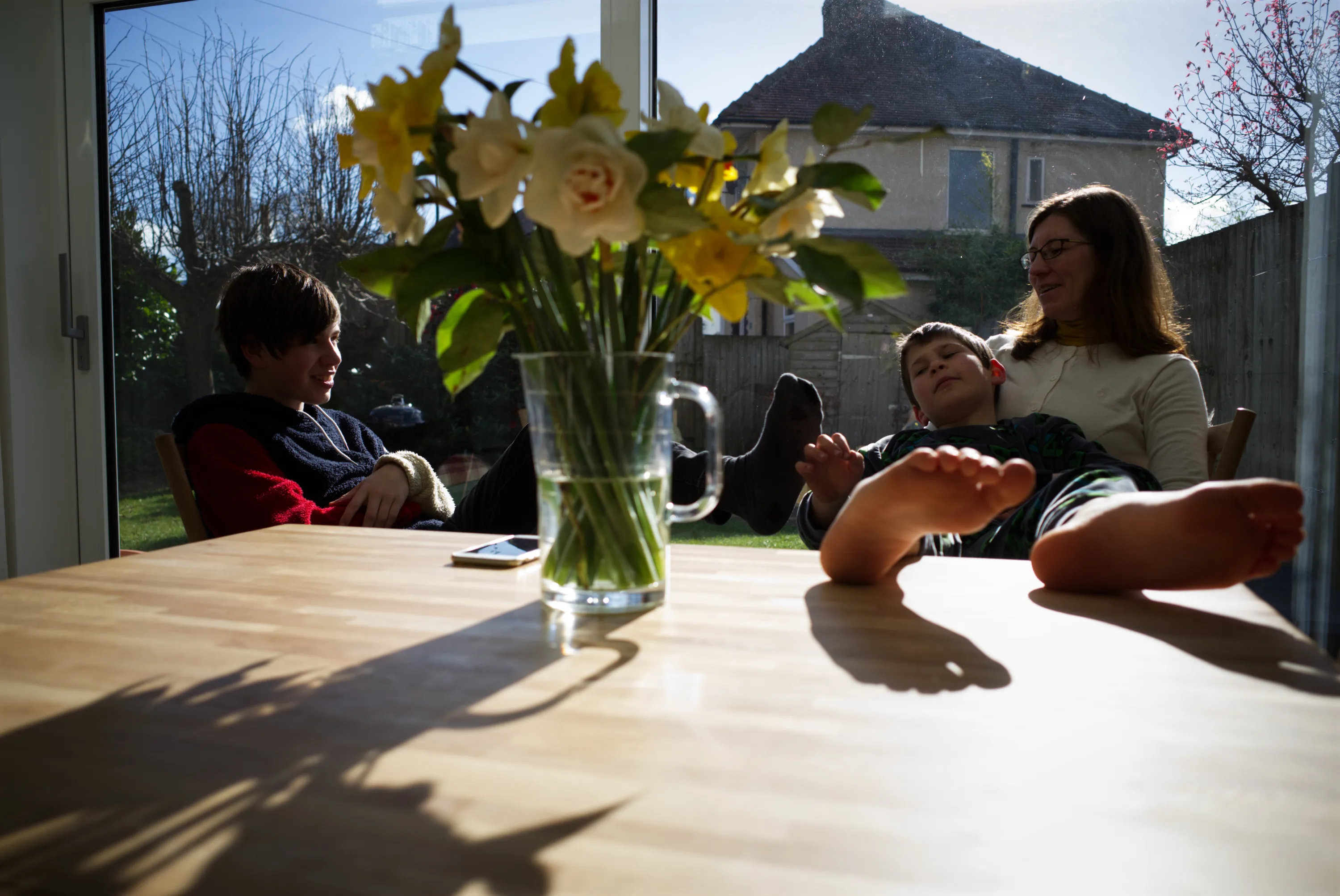 Two children and a woman seated at a dining table with daffodils, glass doors to a garden behind them