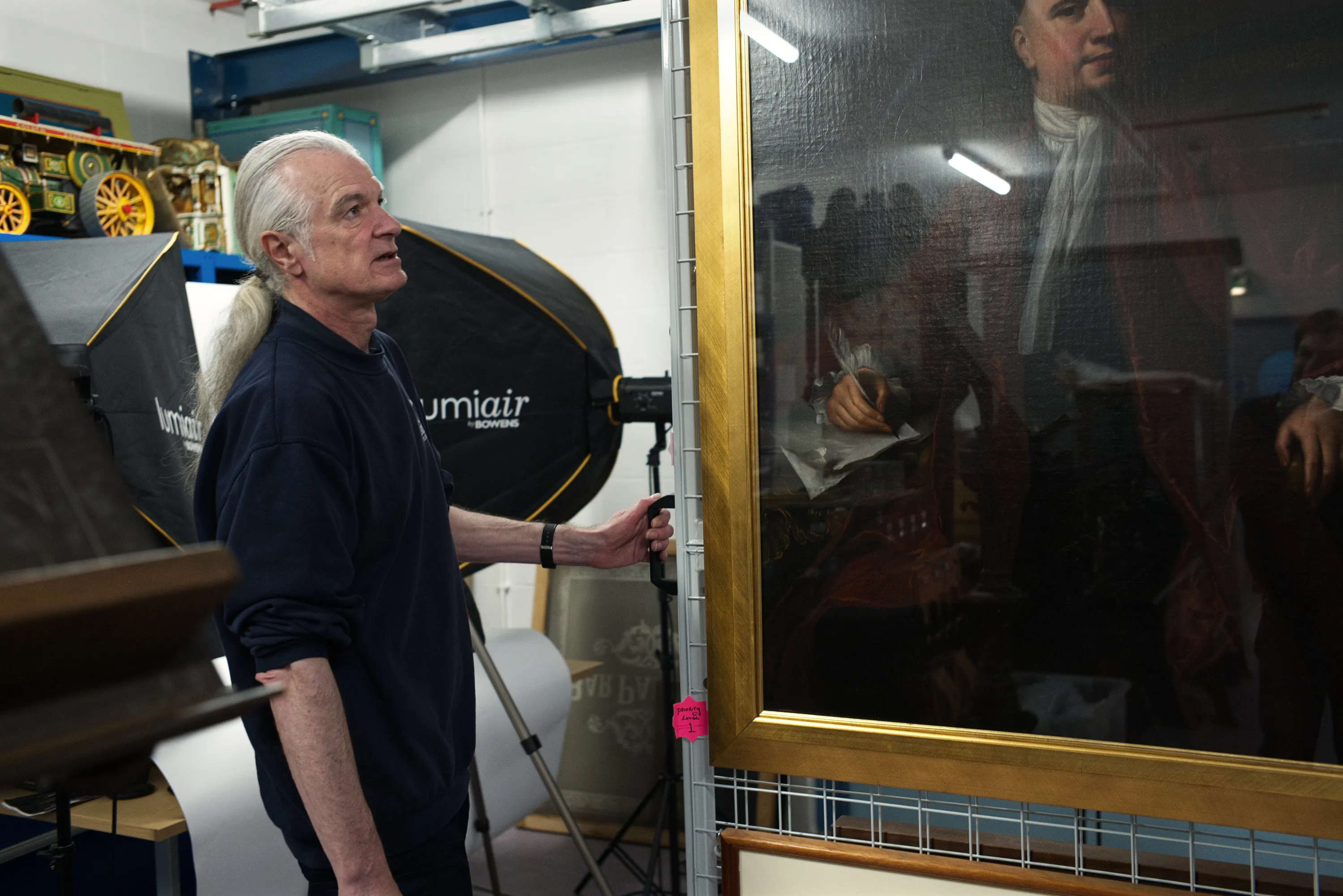 A man with long white hair examines a large gilt-framed portrait painting in a museum storage space, with photography lighting equipment beside him