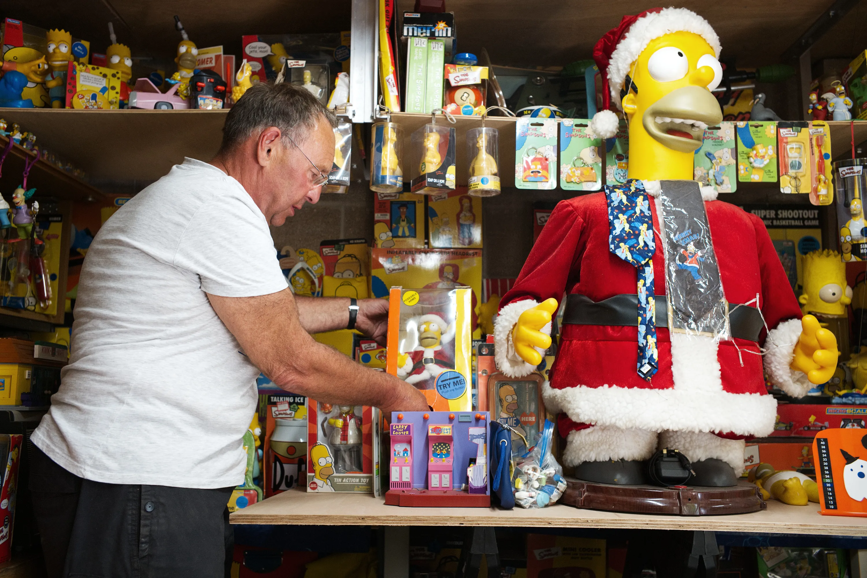 A man arranges Simpsons merchandise and toys at a shelf beside a large Homer Simpson figure dressed as Santa Claus