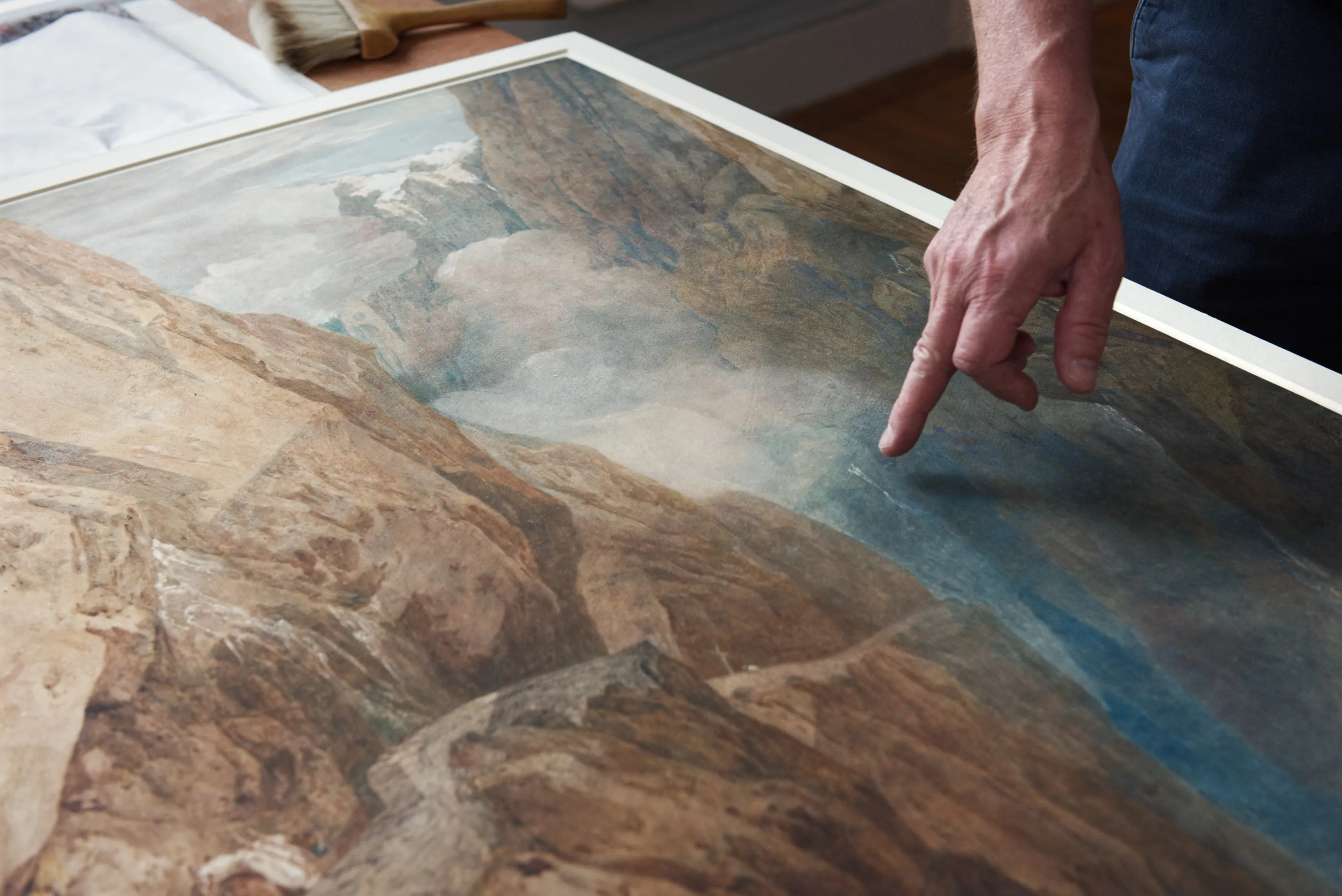 A hand points at the surface of a large landscape watercolour painting lying flat on a table