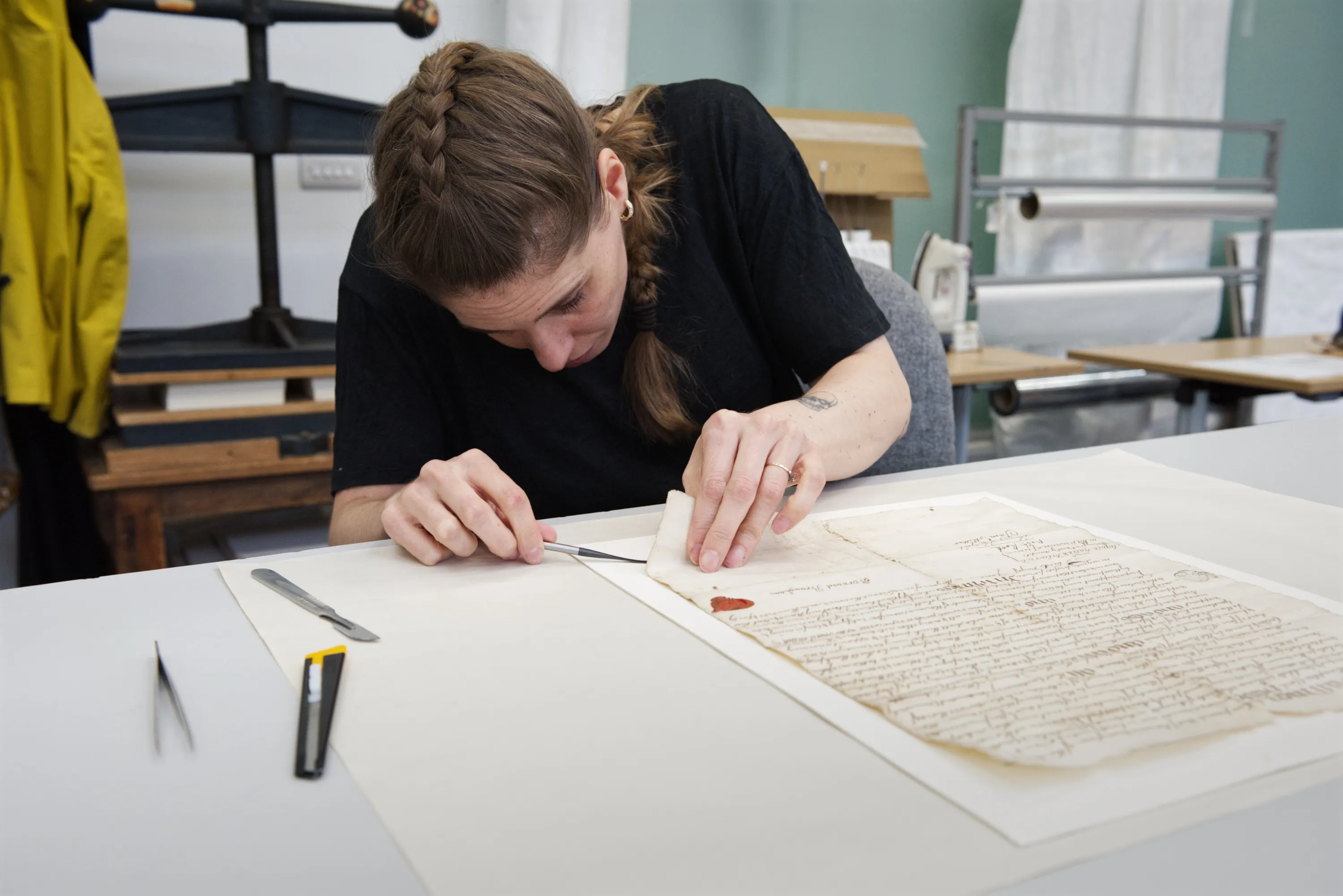A conservator leans over an old manuscript document on a white table, working carefully with tweezers and tools