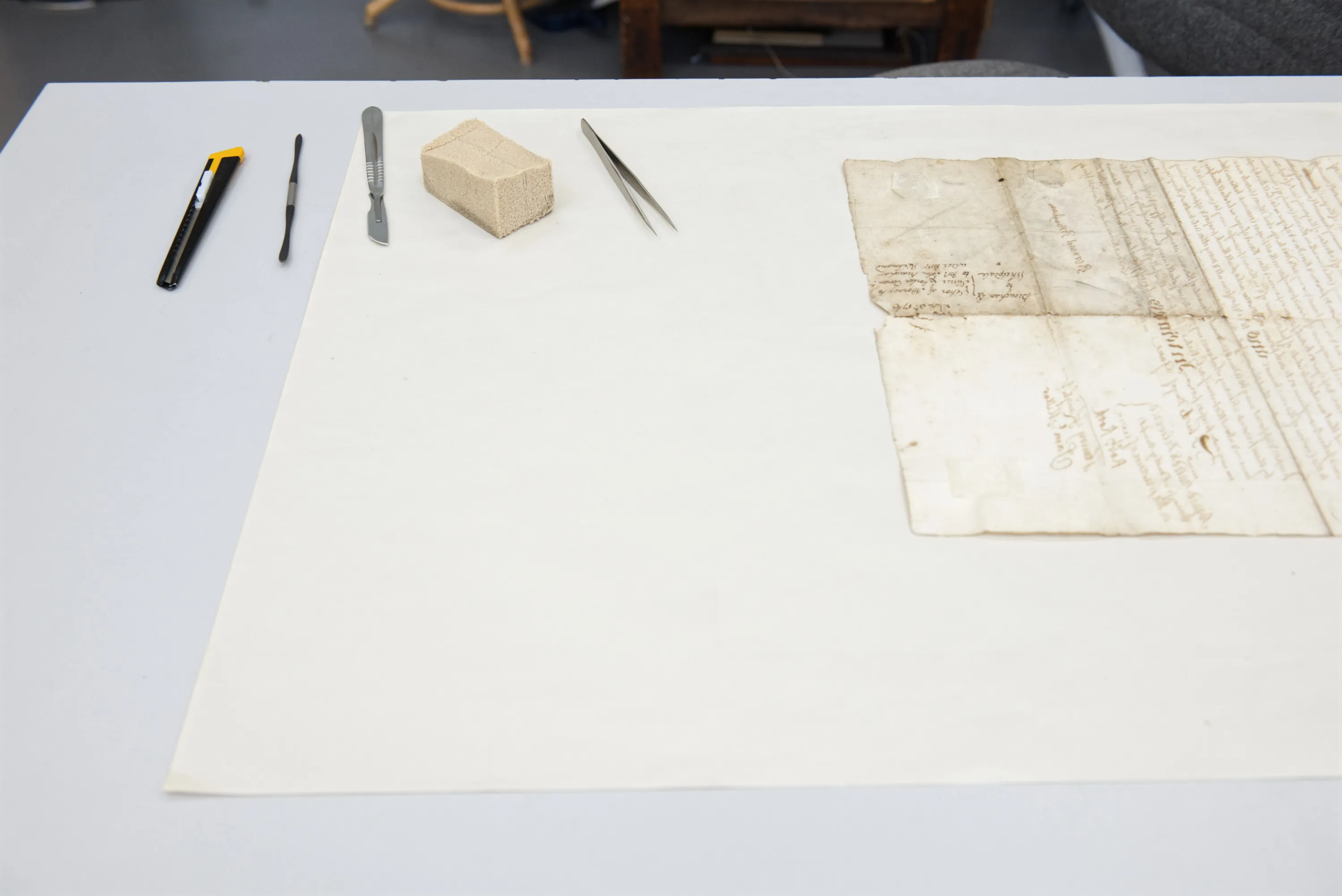 Conservation tools — a scalpel, spatula, eraser block, and tweezers — laid out beside an aged manuscript on a white table