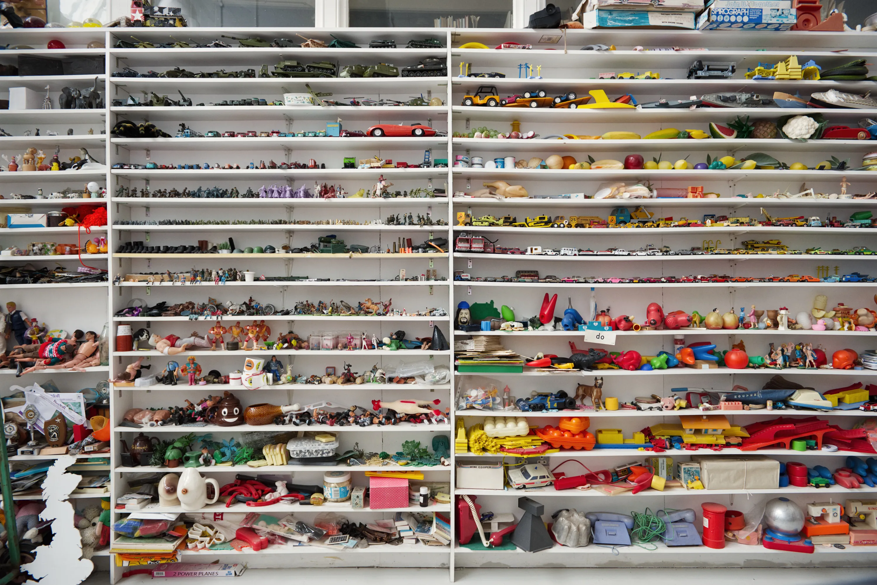 Floor-to-ceiling white shelving units packed with hundreds of small plastic toys, figurines, and miscellaneous objects in every colour