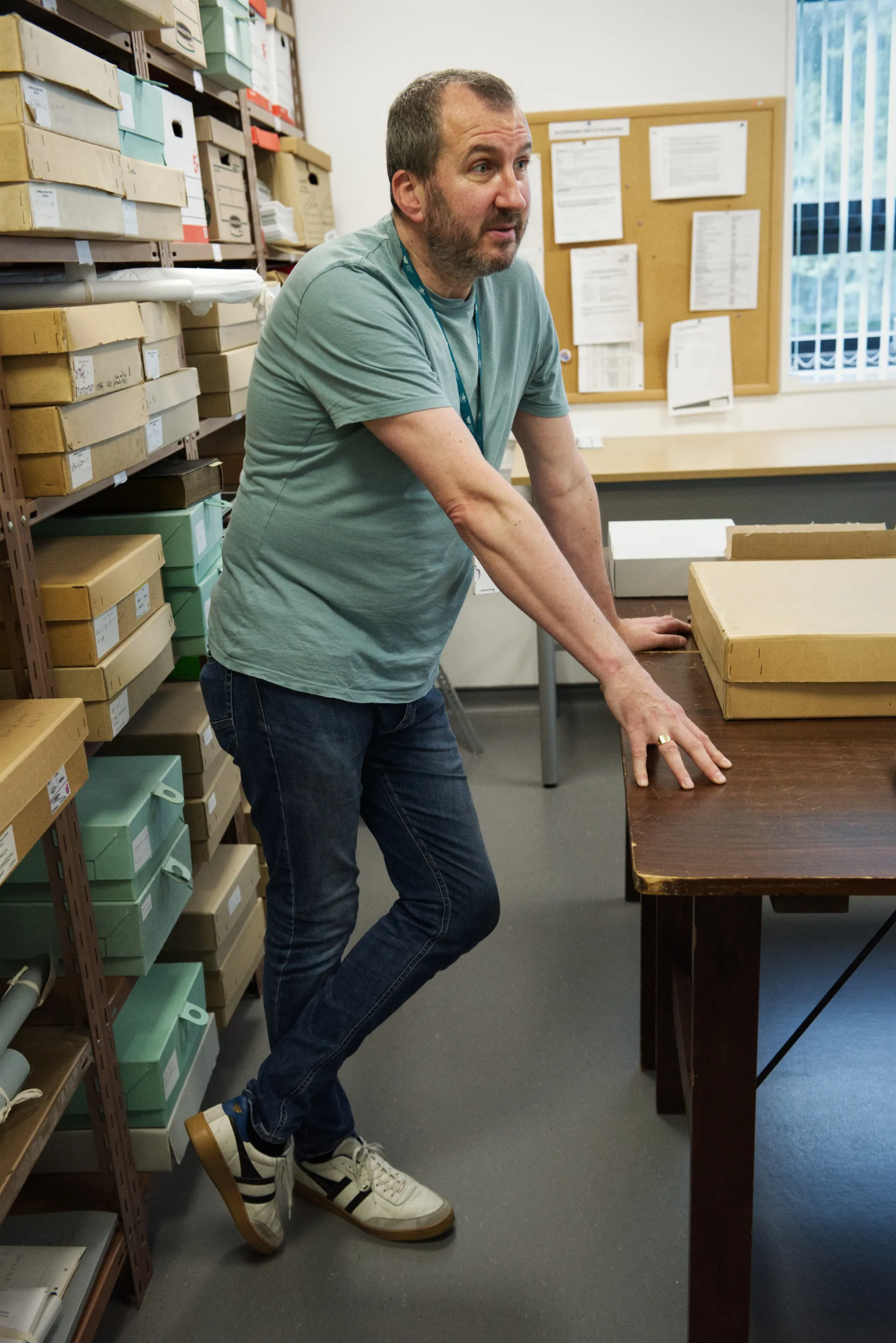 A man in a teal t-shirt stands in a dense archive storeroom lined with flat archival boxes on metal shelving
