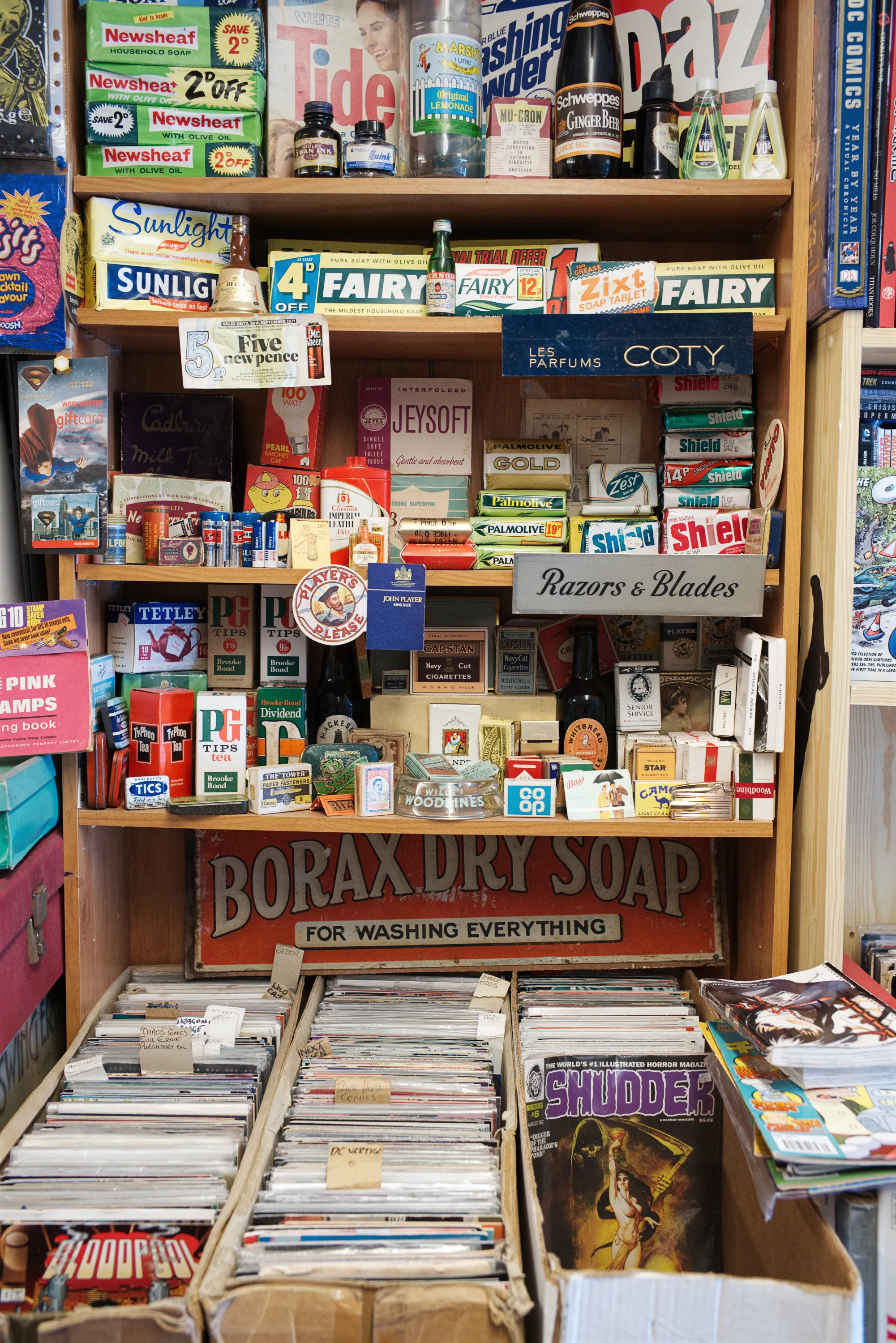 A shelving unit crowded with vintage household product packaging — soap powder boxes, razor blades, and other branded tins and packets — with boxes of comics below