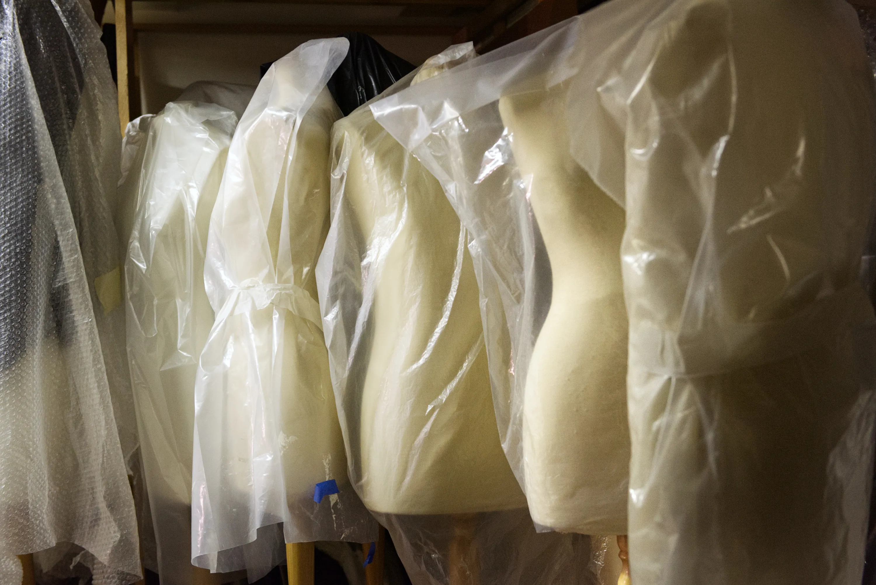 A row of dressmaker's mannequin torsos wrapped in translucent plastic bags hanging in a dark storage space