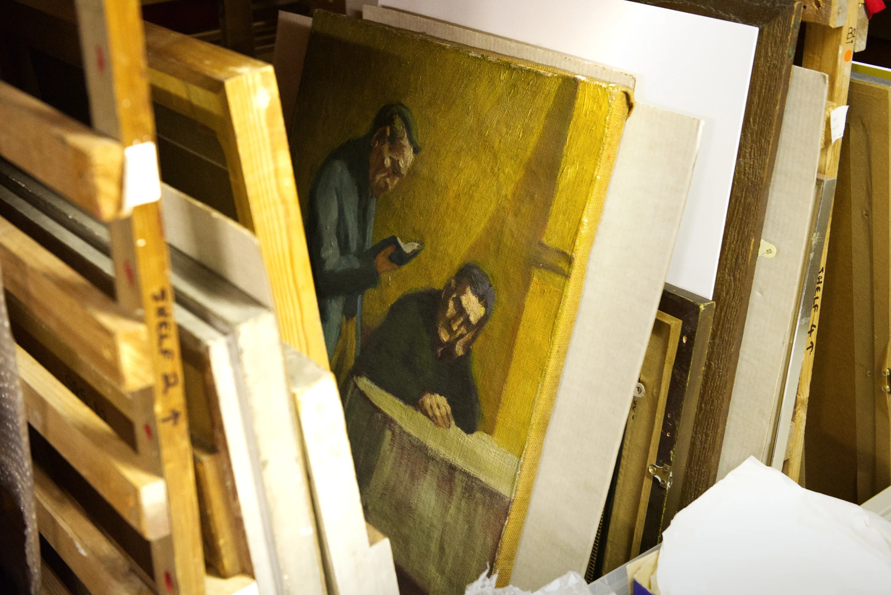 Unframed paintings stacked on wooden racks in an art store; the foremost shows two figures under a yellow light