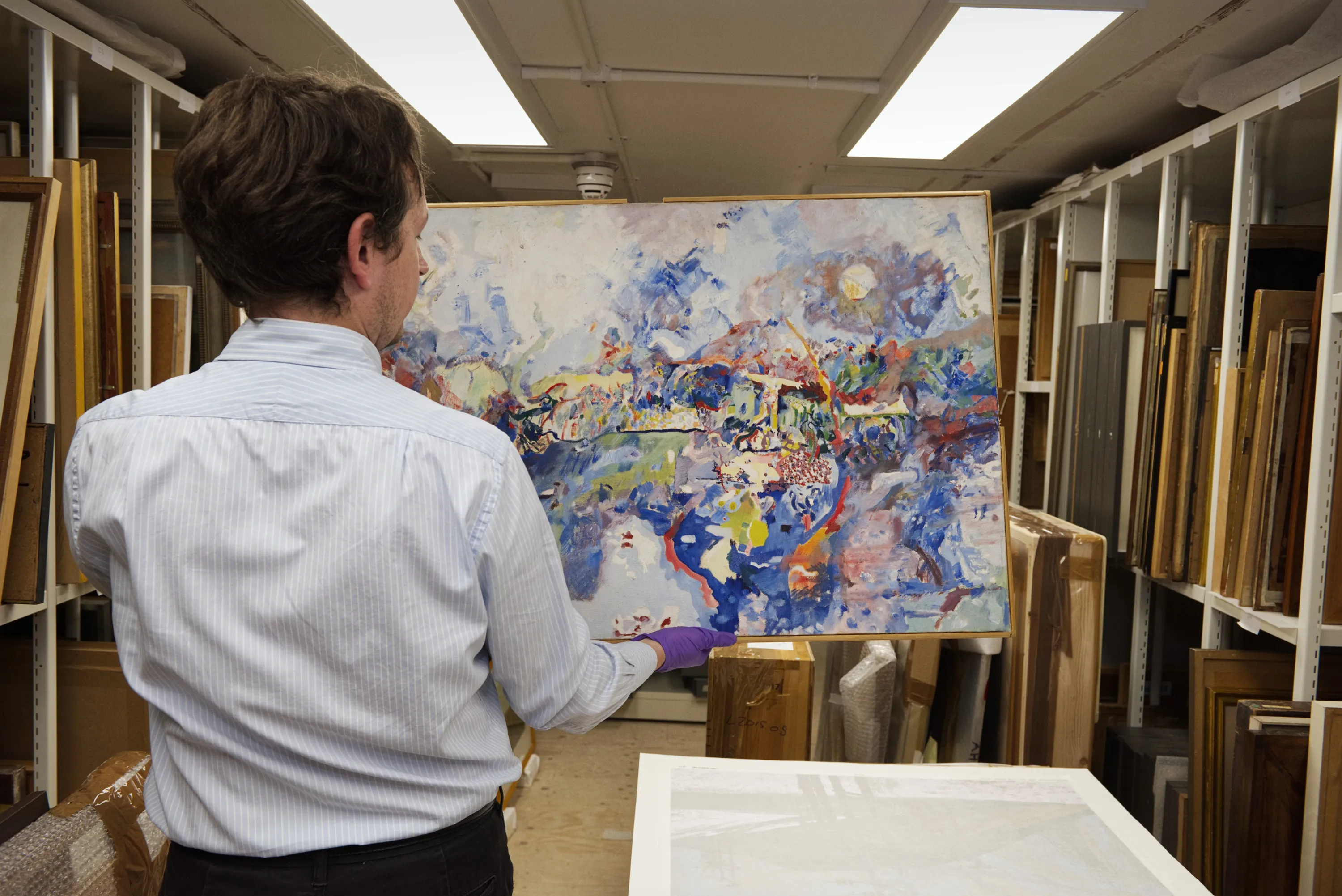 A curator holds an abstract painting in a gallery art store, surrounded by racks of framed and unframed works