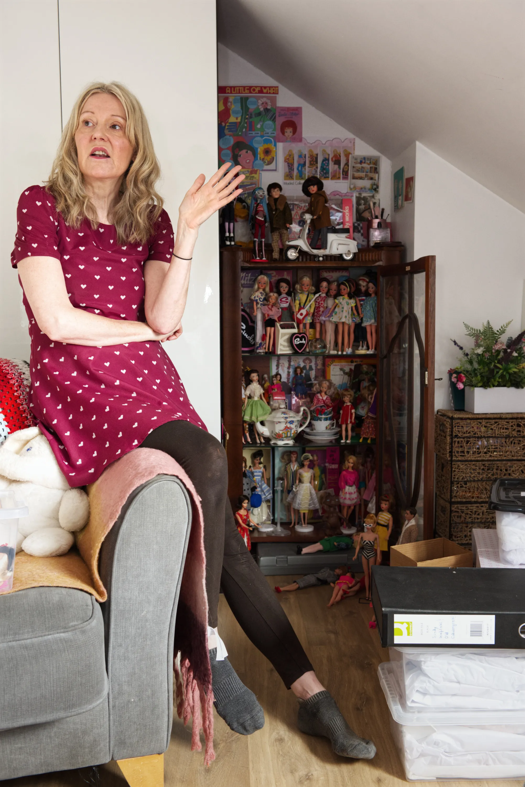 A woman sits in an armchair gesturing expressively, with a tall glass cabinet of Barbie dolls and doll-related memorabilia behind her