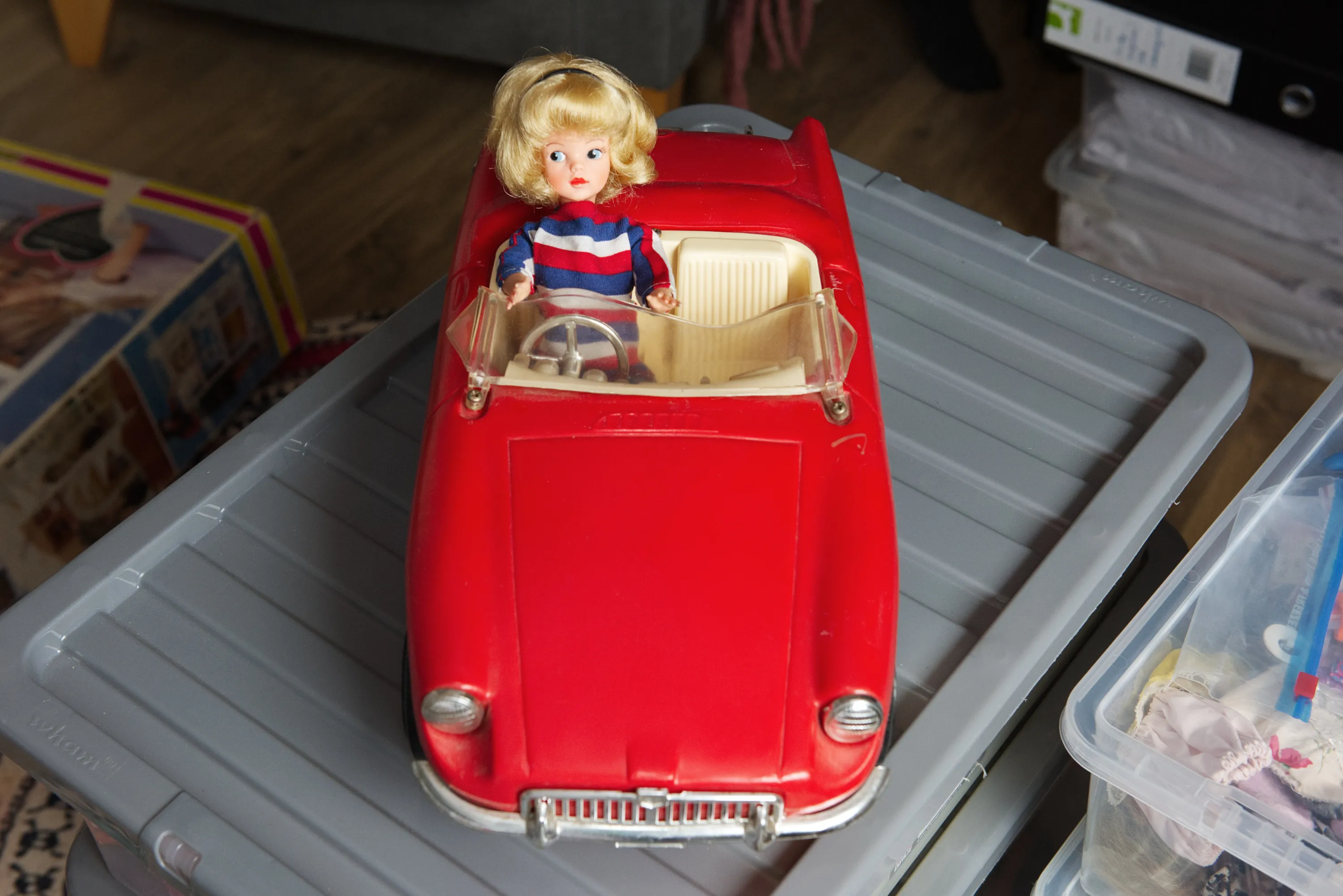 A vintage blonde doll seated in a red toy convertible car on a plastic storage box