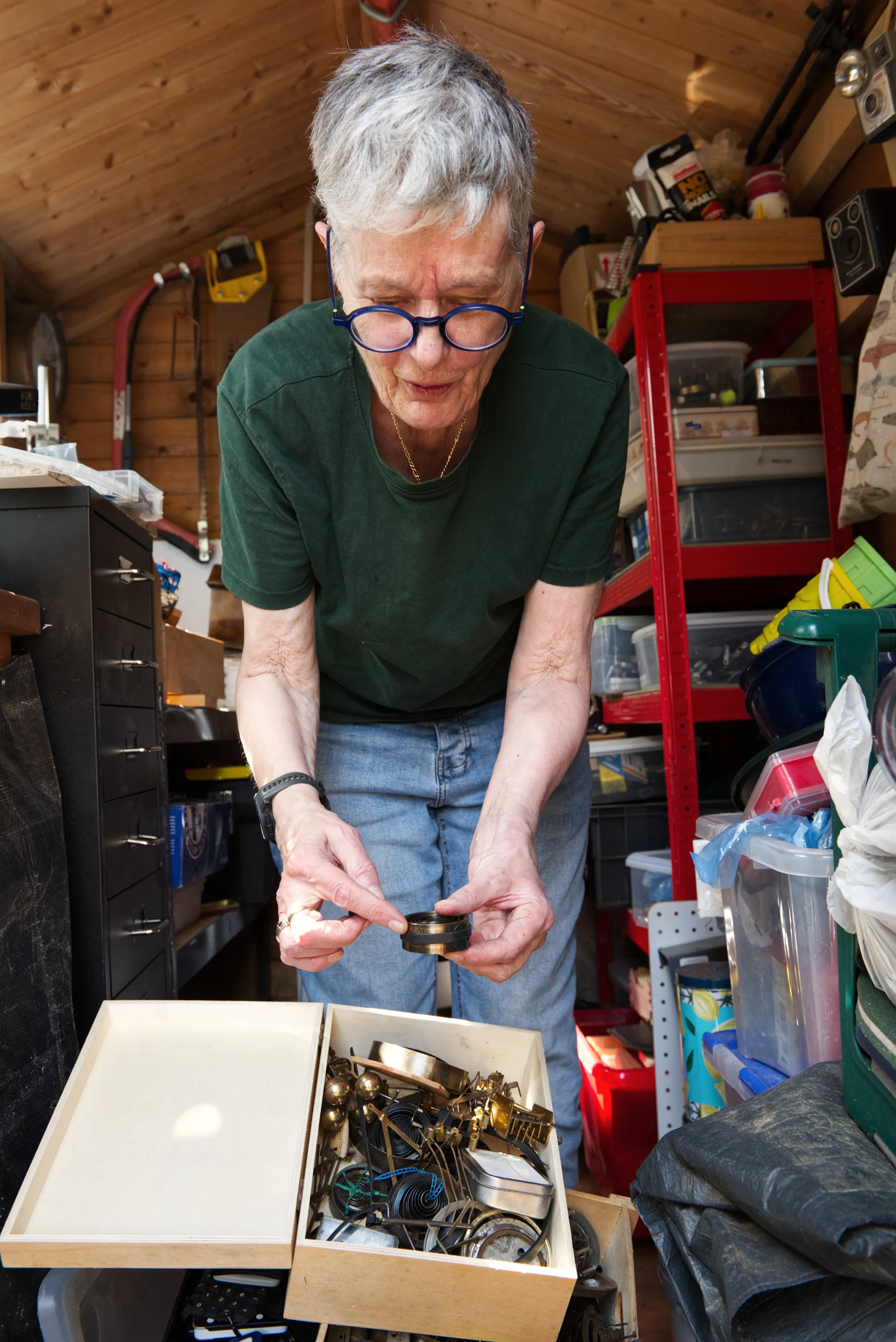 An elderly woman examines a small object from a box of mixed brass hardware and watch parts in a cluttered garden shed