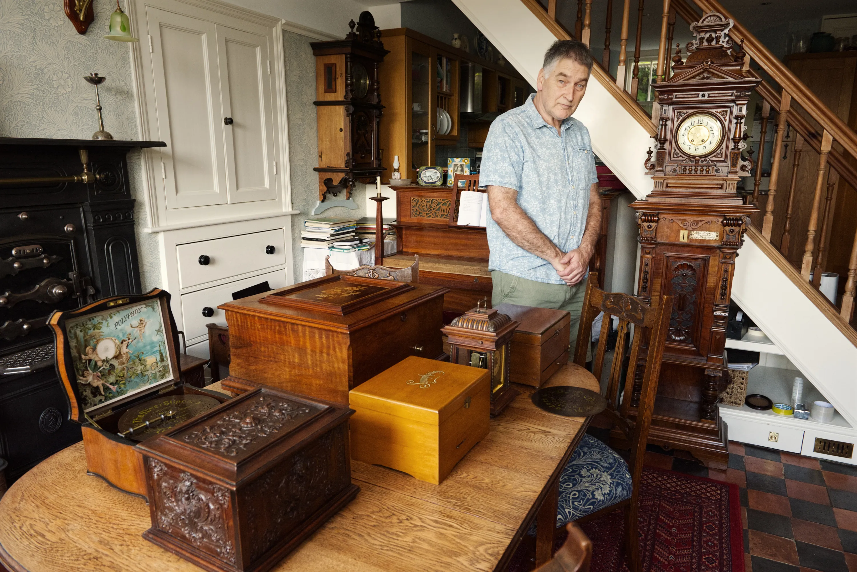 A man stands in his sitting room surrounded by a collection of antique wooden music boxes of various sizes, with a tall ornate clock against the wall