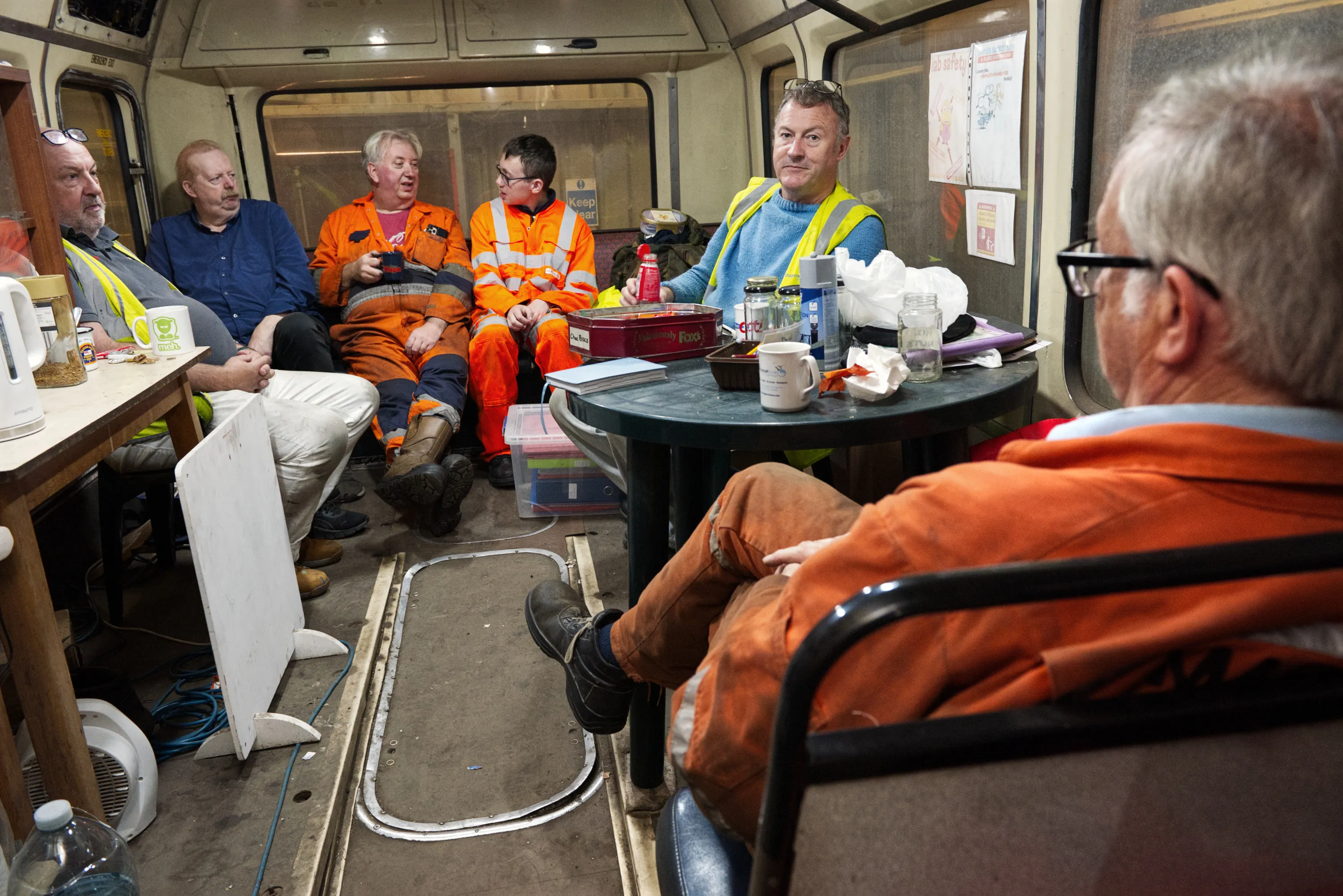 Five men in work clothes and high-visibility vests sitting inside a bus, talking during a break