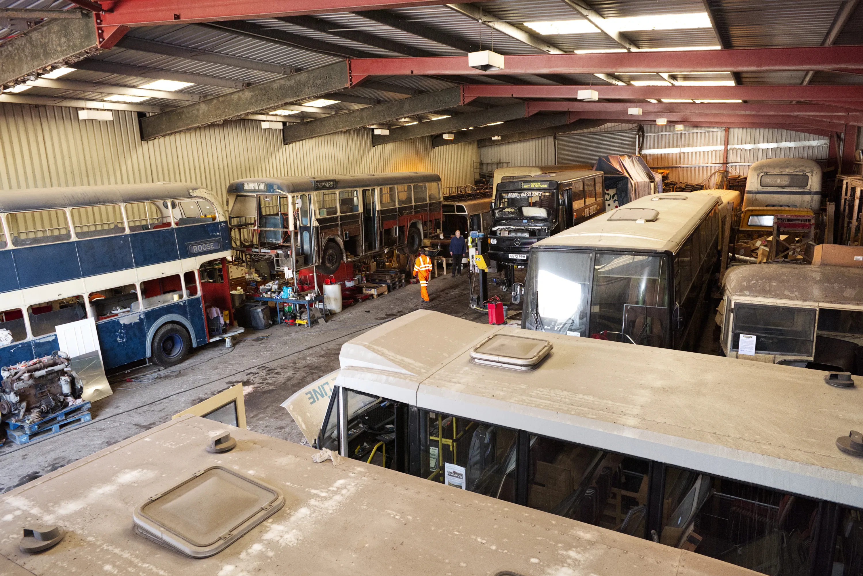 An elevated view inside a large industrial workshop with several vintage double-decker and single-decker buses in various states of restoration