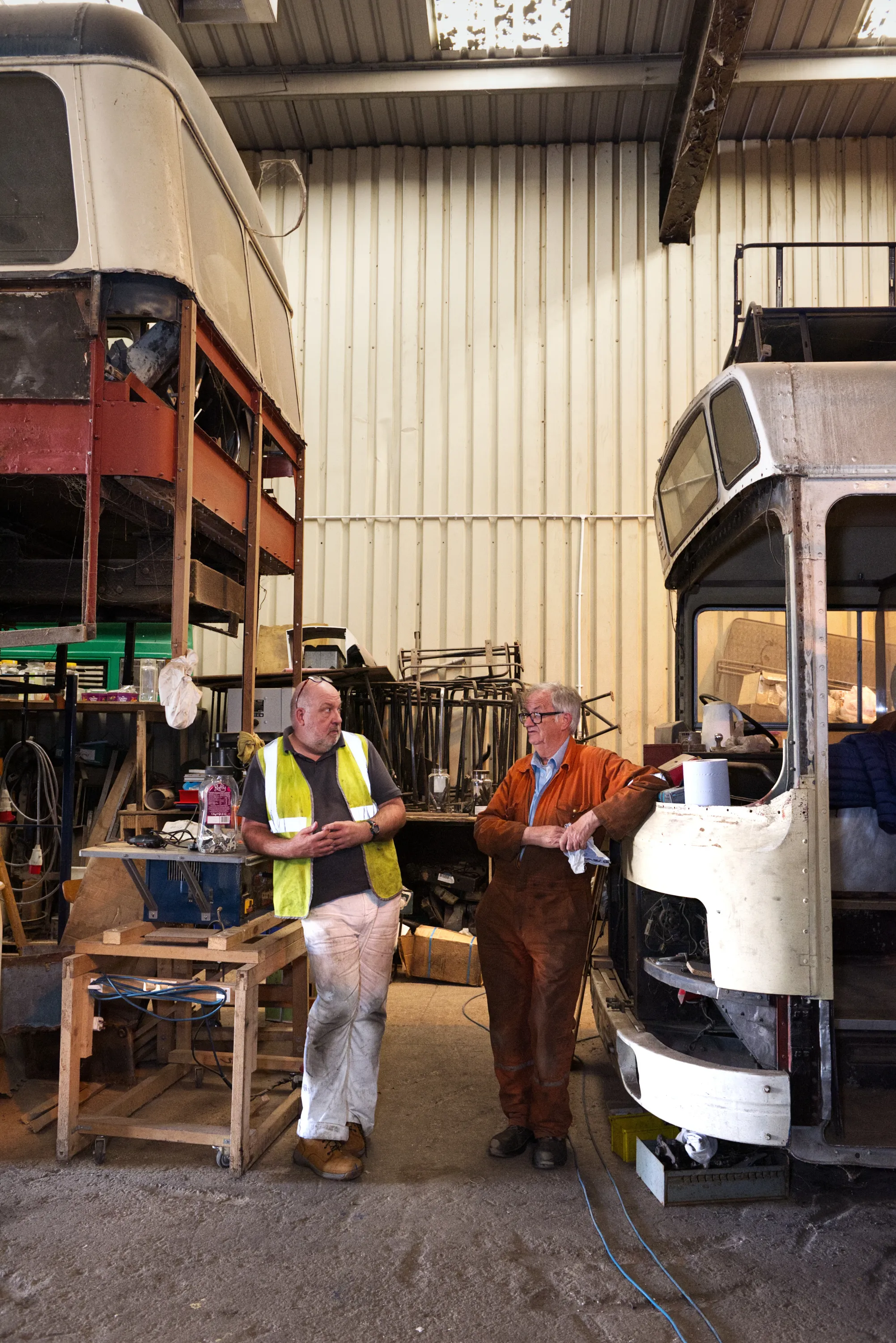 Two men stand talking beside a partially stripped vintage bus body in a workshop, one in a hi-vis vest, the other in a brown work coat