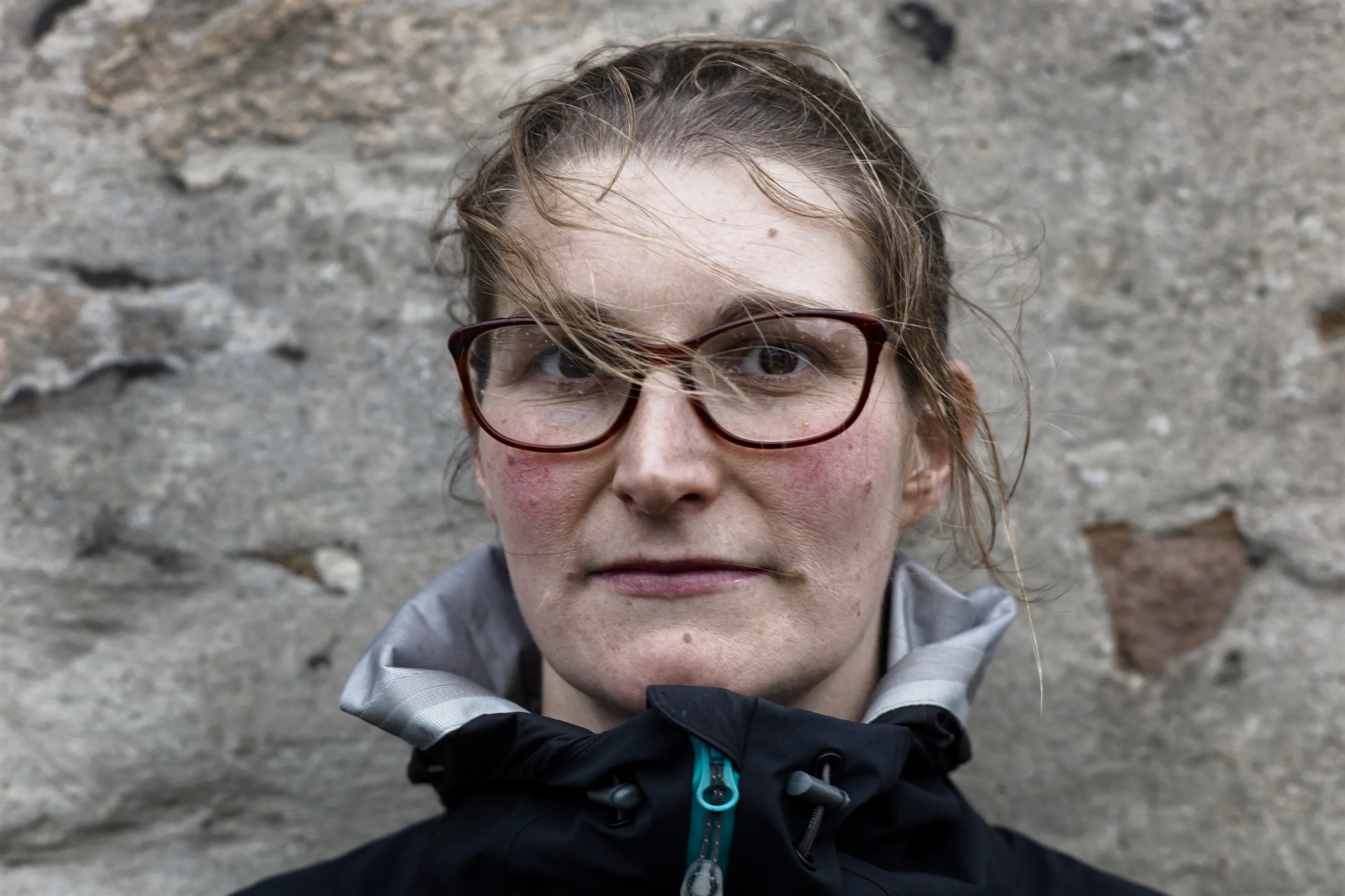 Close portrait of a woman with red-framed glasses against a stone wall