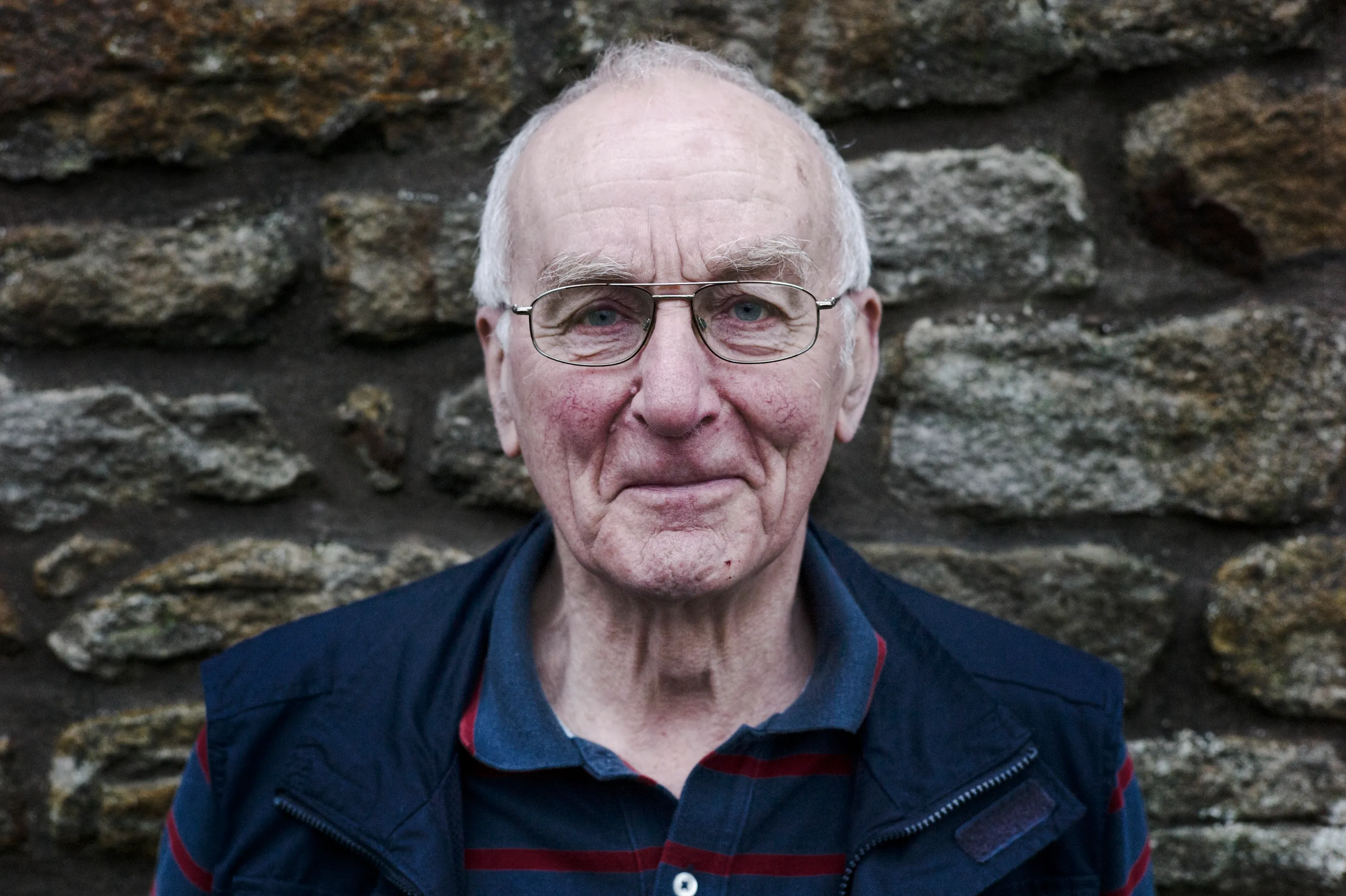 Close portrait of a man with wire-framed glasses against a stone wall