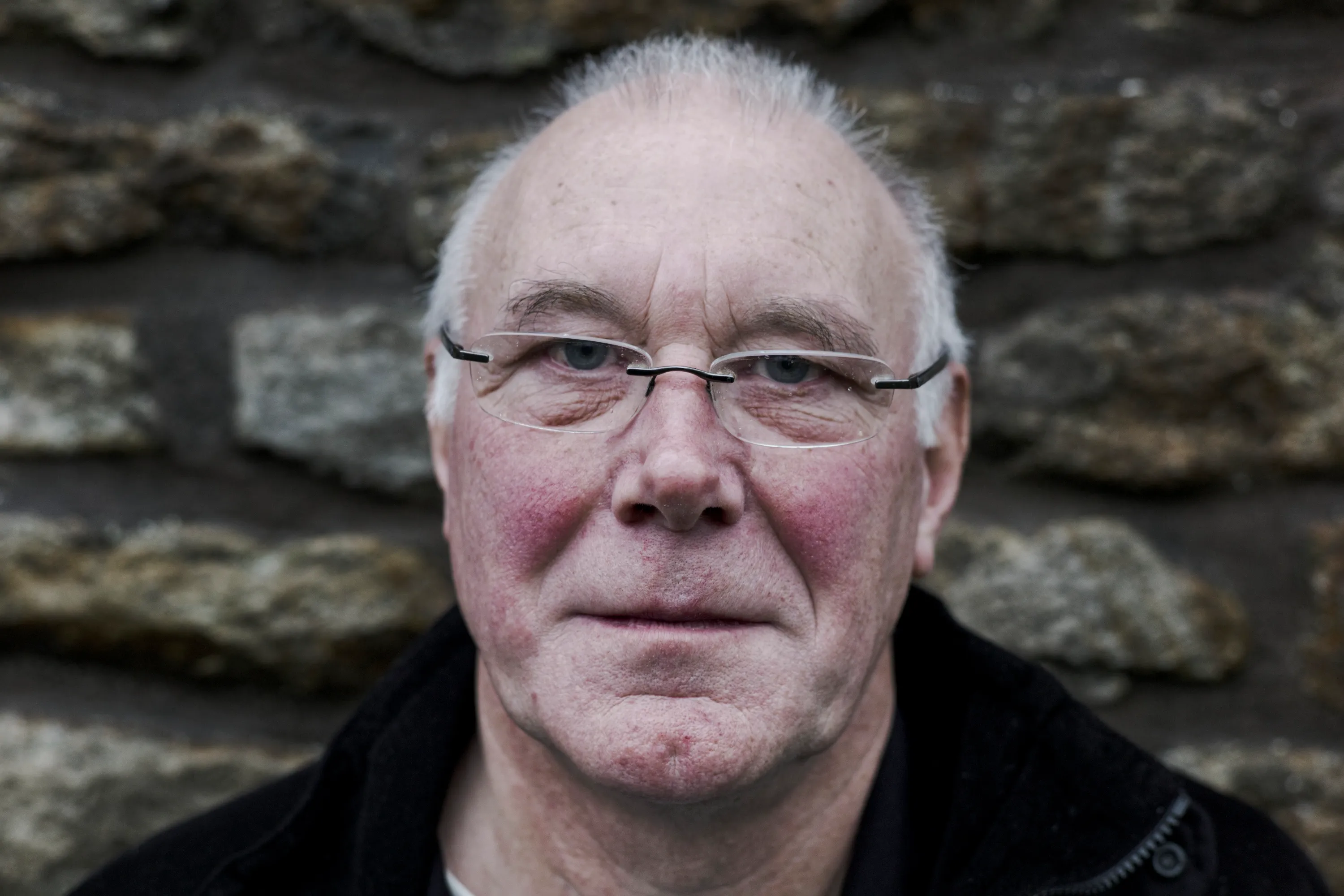 Close portrait of a man with rimless glasses against a stone wall