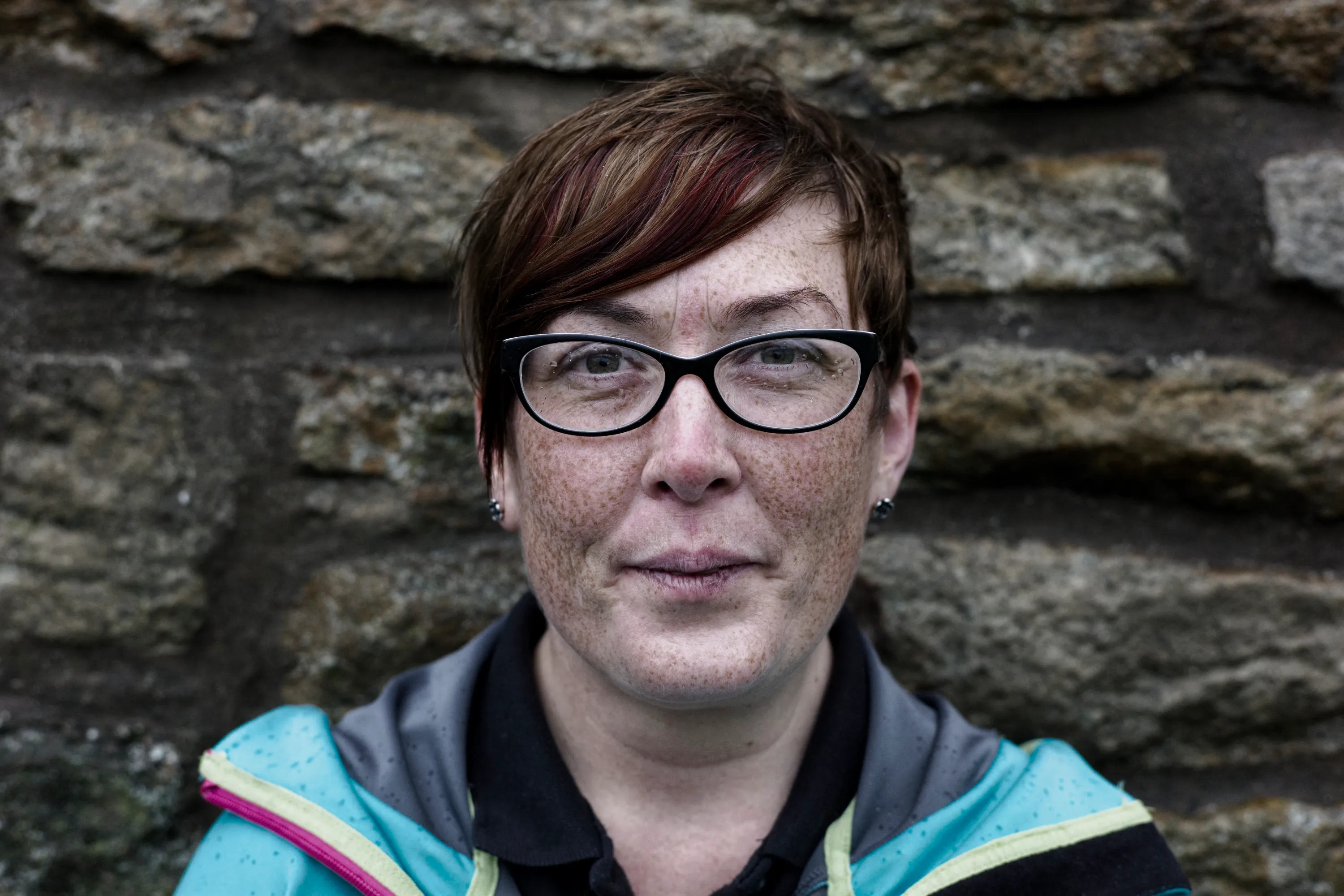 Close portrait of a woman with short red-brown hair and black-framed glasses against a stone wall