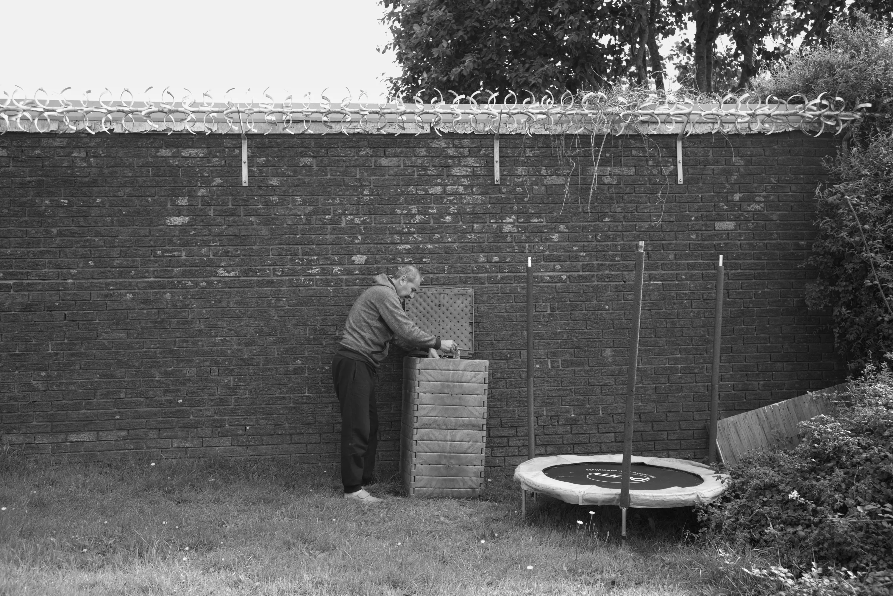 Man tending a wooden compost bin against a brick wall with a small trampoline nearby