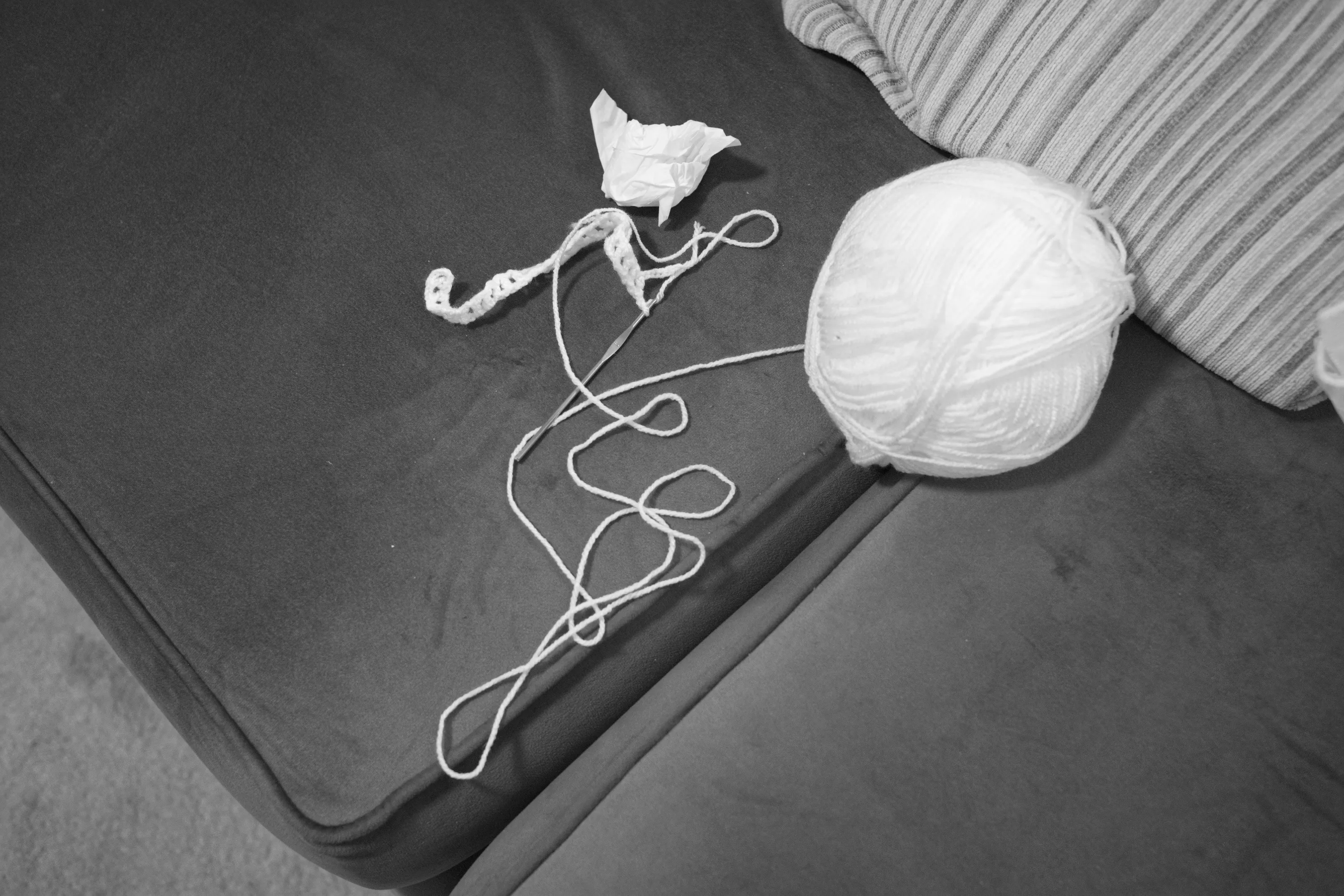 Ball of white yarn and a small piece of crochet work on a grey sofa