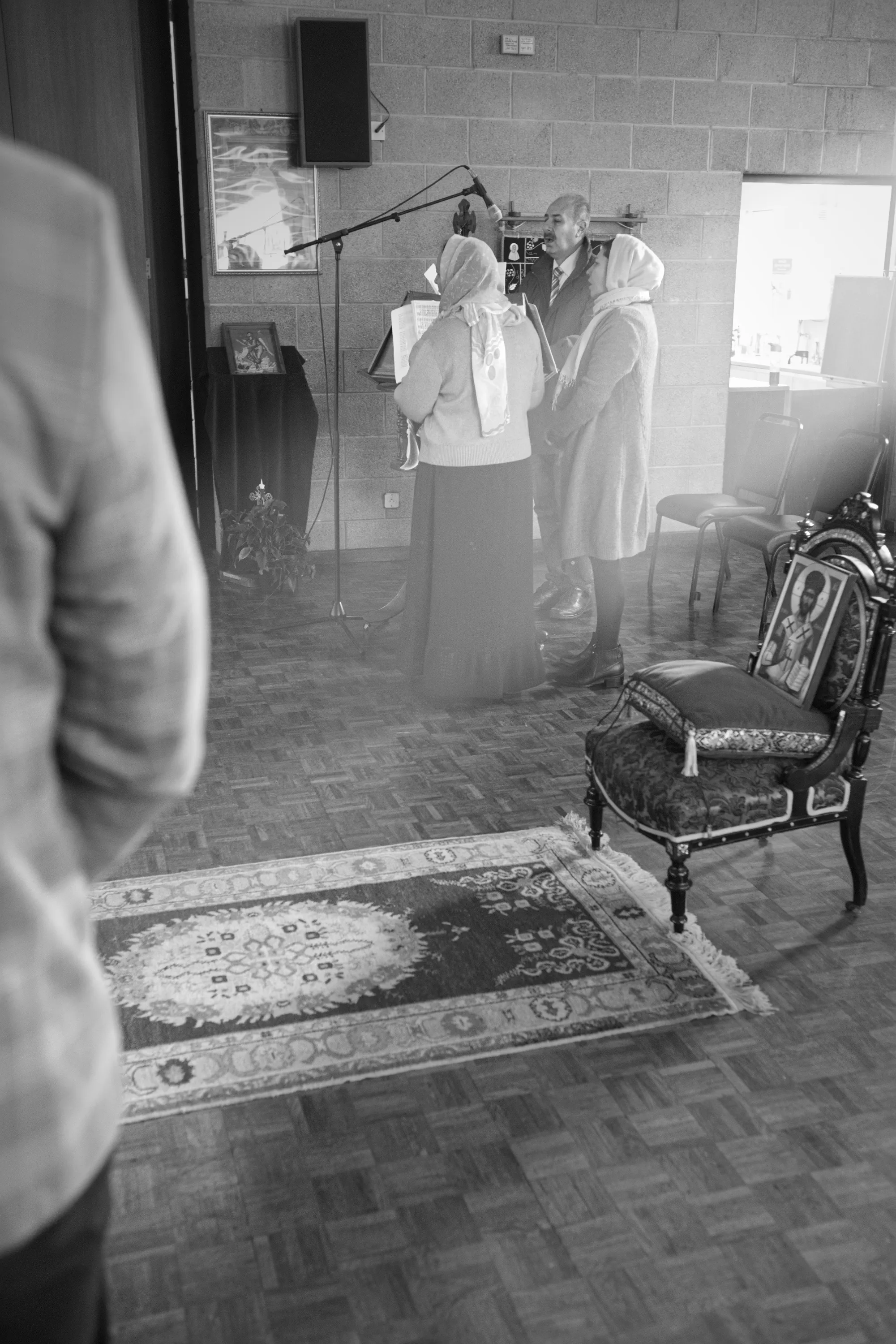 Two women and a man standing near a microphone stand in a bare-walled hall, an ornate chair in the foreground