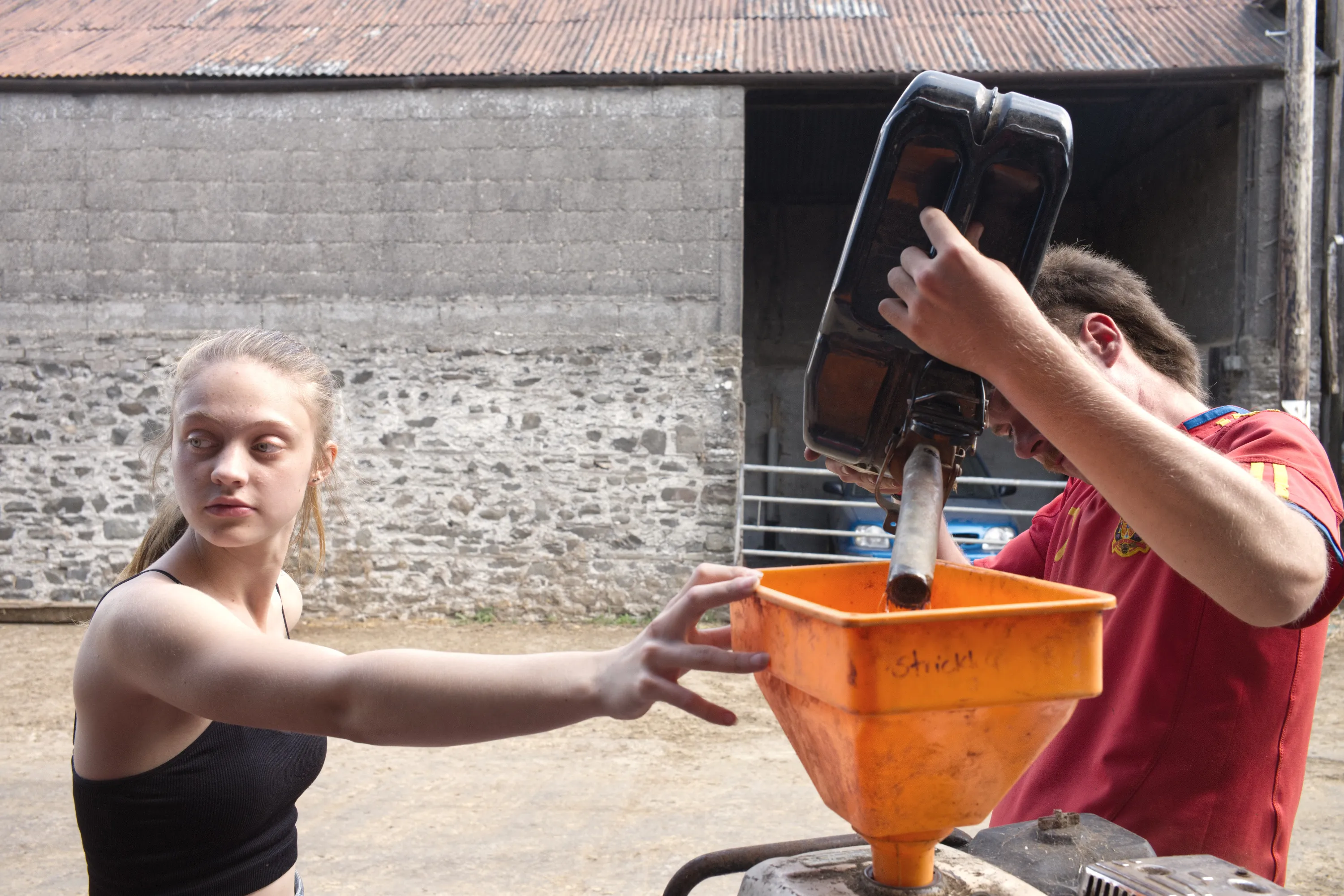 Young woman and young man refilling machinery using a large orange funnel in a farmyard