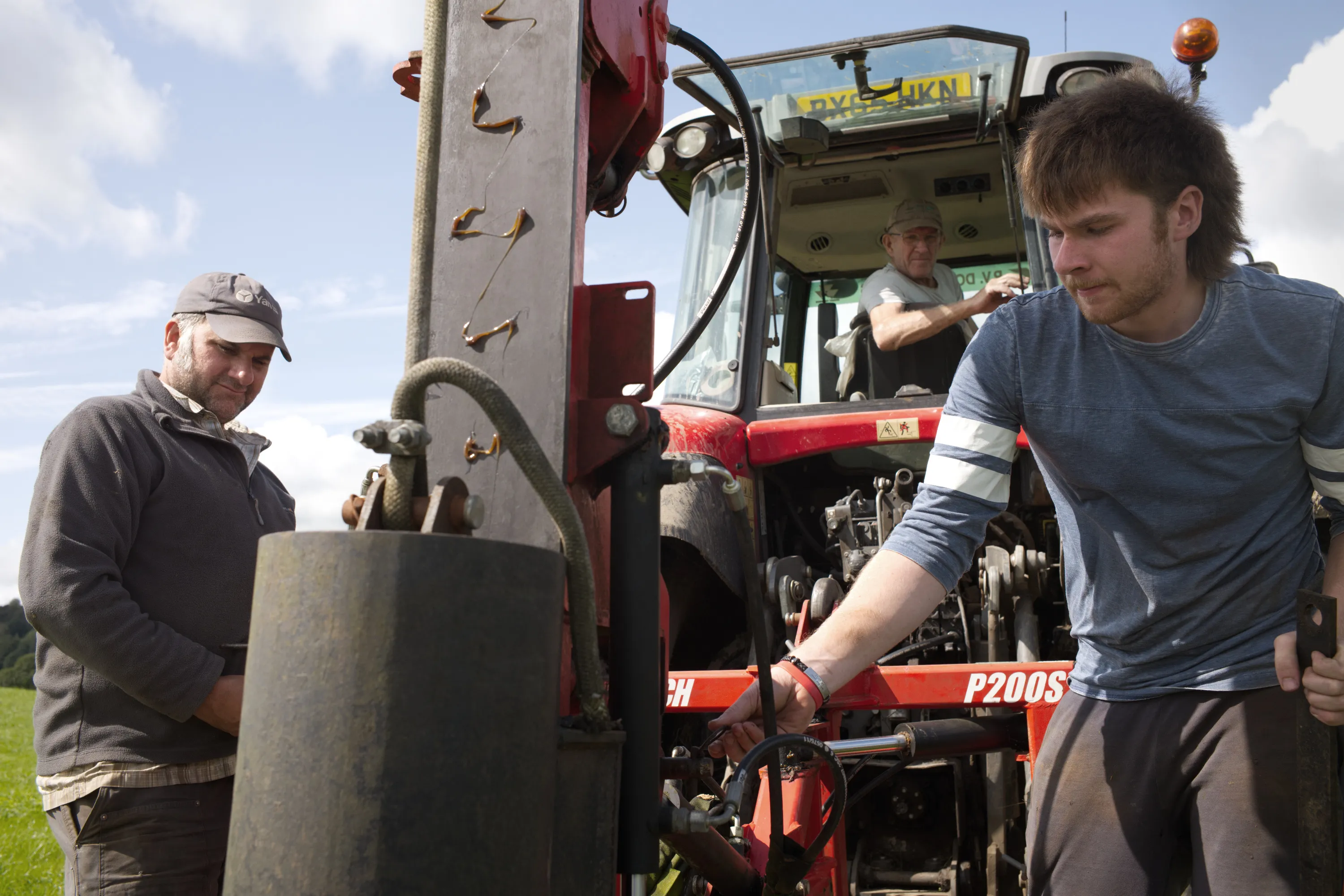 Three men working around a red tractor with a post-driver attachment in a field