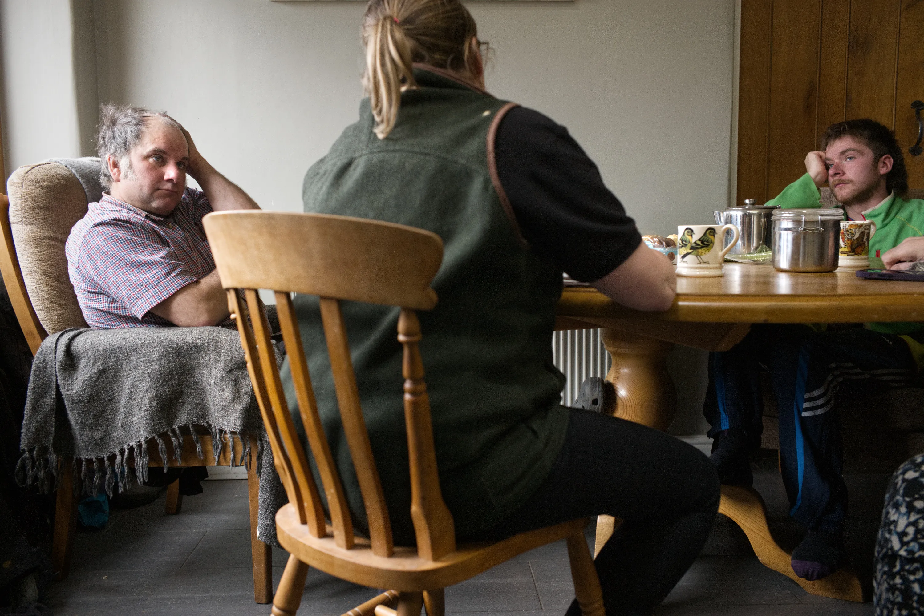 Three people seated around a kitchen table with mugs, engaged in conversation