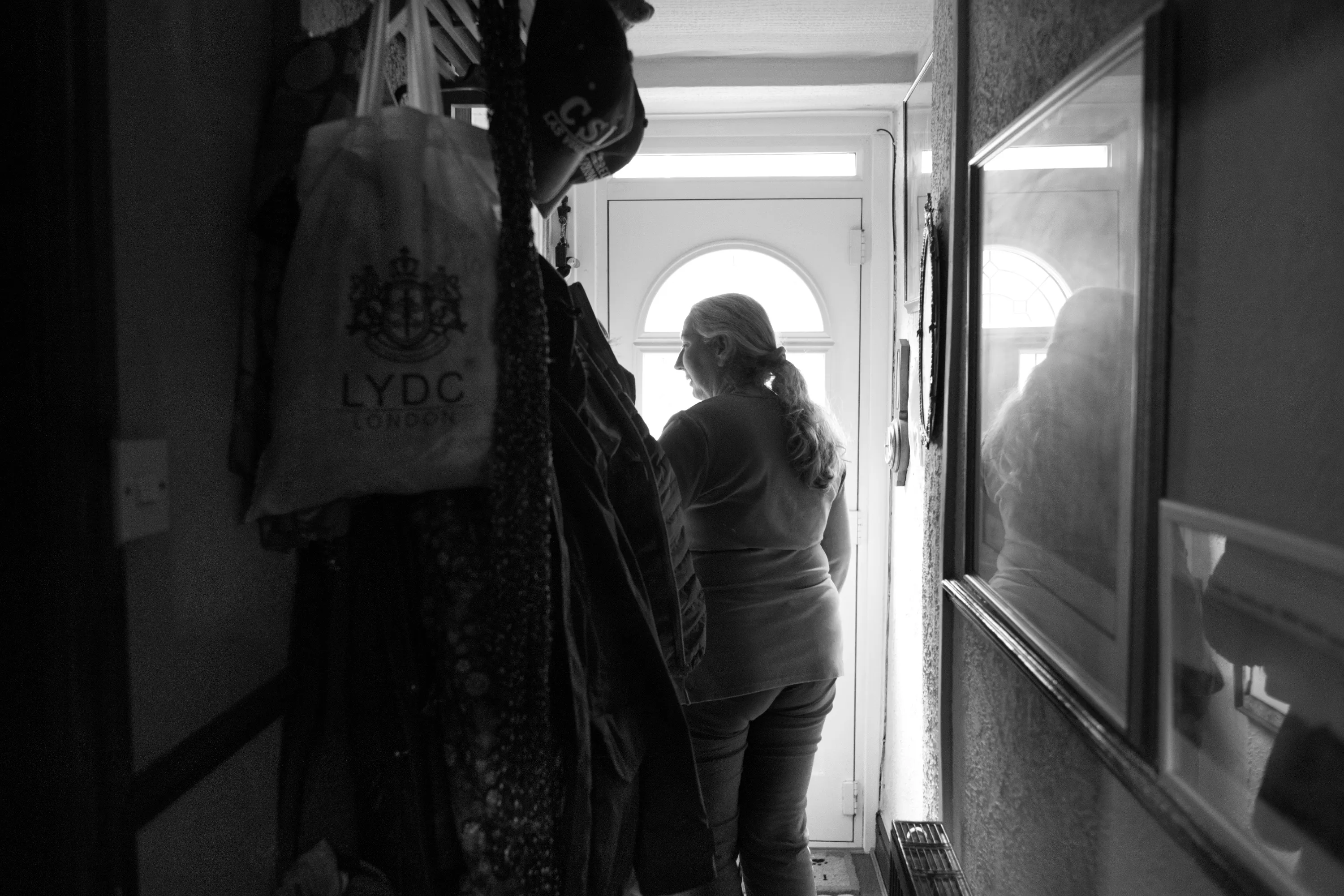 A woman's silhouette from behind in a narrow hallway, her profile lit by light through the arched glazing of the front door, coats and a tote bag hanging to the left