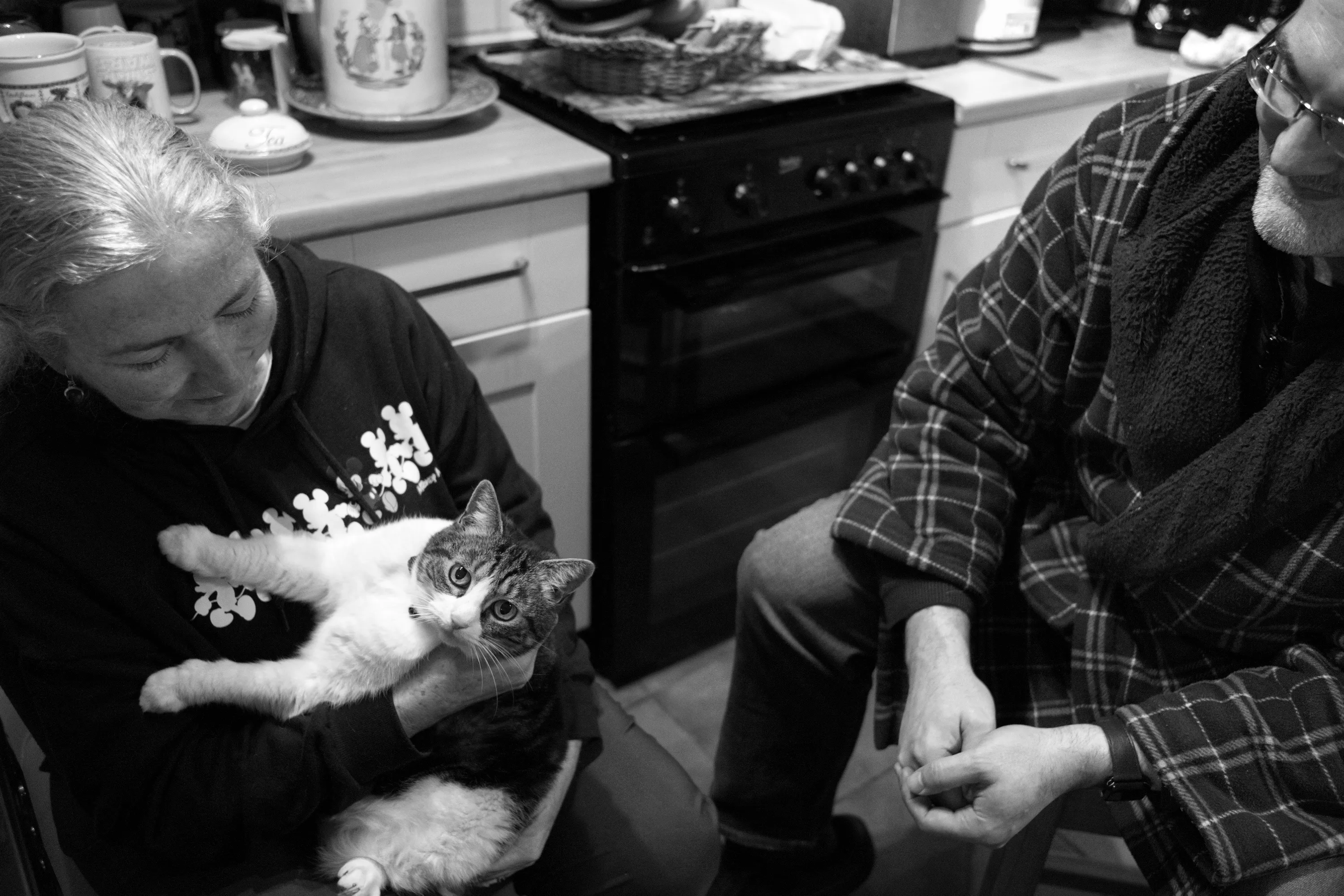 A woman seated in the kitchen holding a Manx cat in her arms, with a man sitting beside her