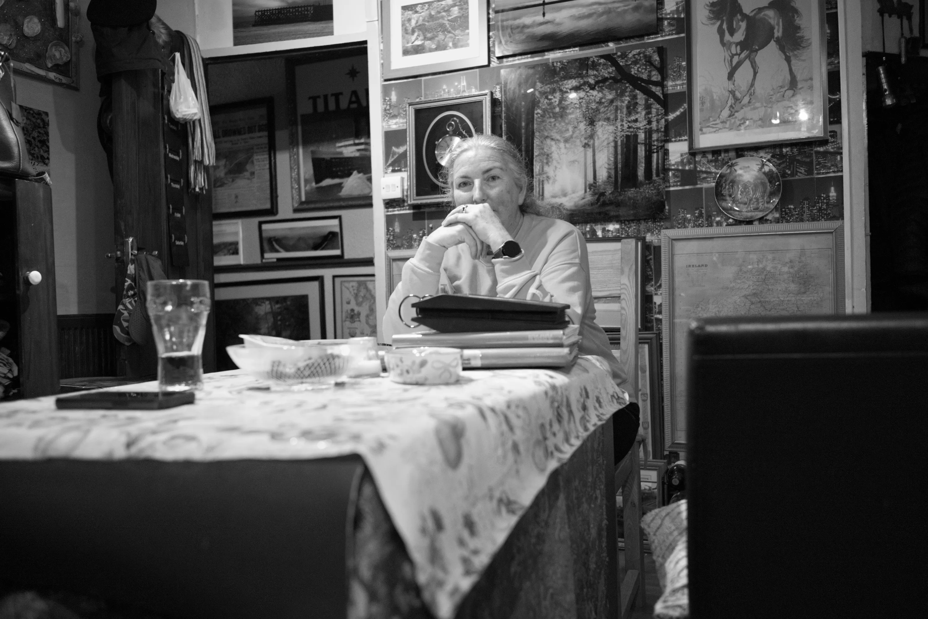 A woman seated alone at the dining table, chin resting on her hands, surrounded by a wall covered in framed pictures and prints