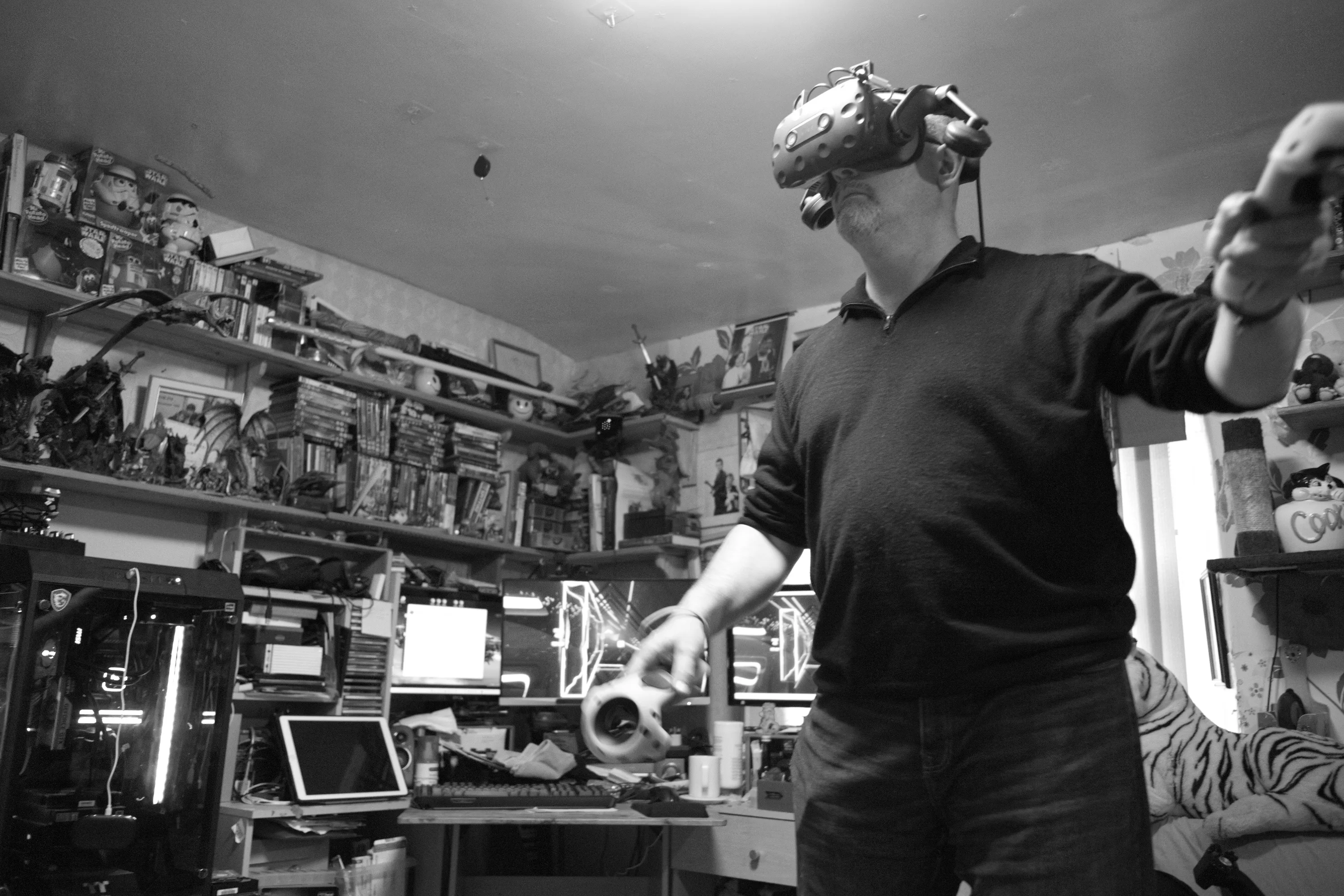 A man standing in a cluttered room wearing a VR headset and holding controllers, arms outstretched, shelves of games and collectibles behind him