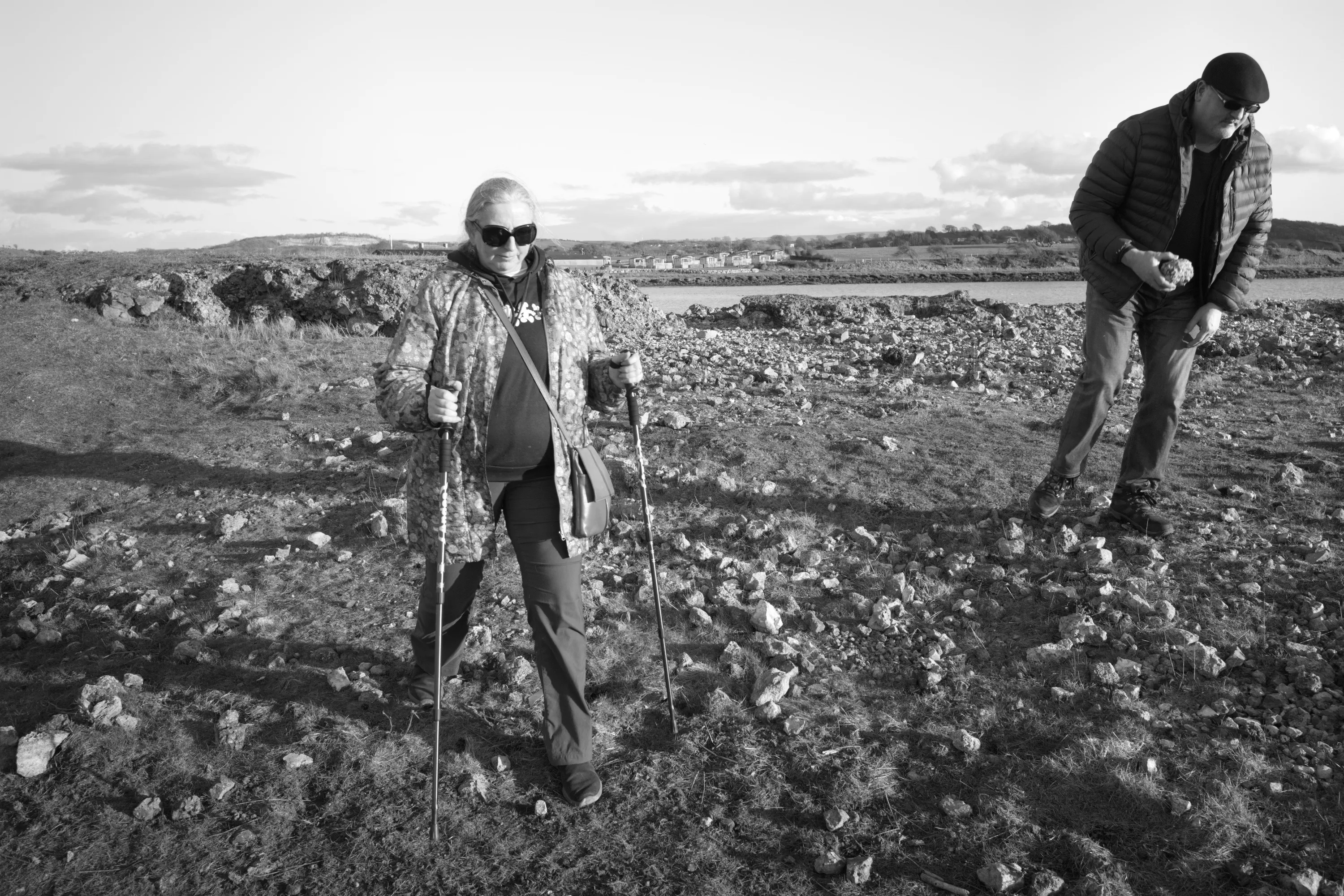 A woman smiling and walking on open rocky ground with two walking sticks, a man bending down to pick something up in the background