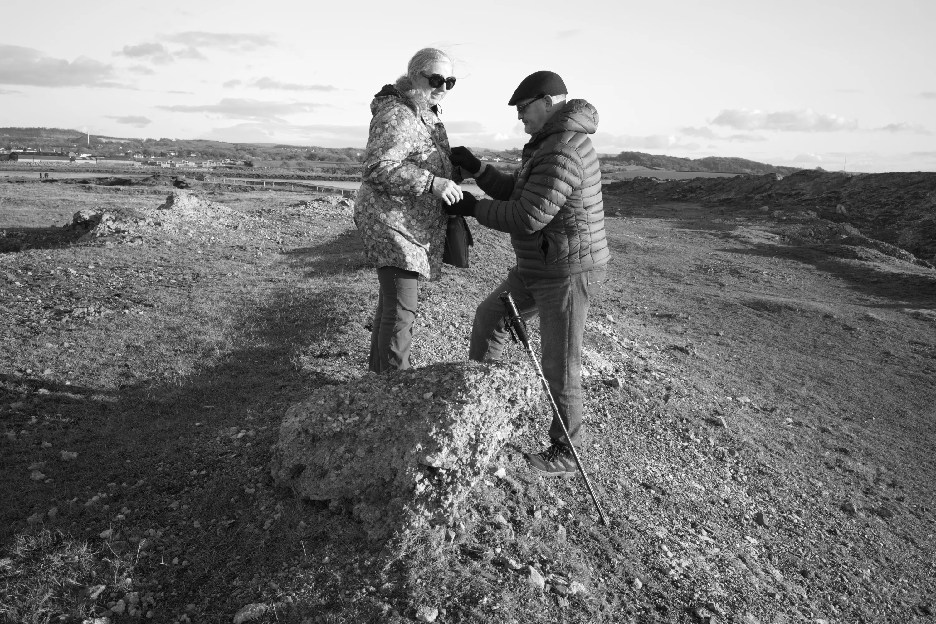 A man steadying a woman on a rocky outcrop outdoors, both dressed in winter coats, a wide flat landscape behind them