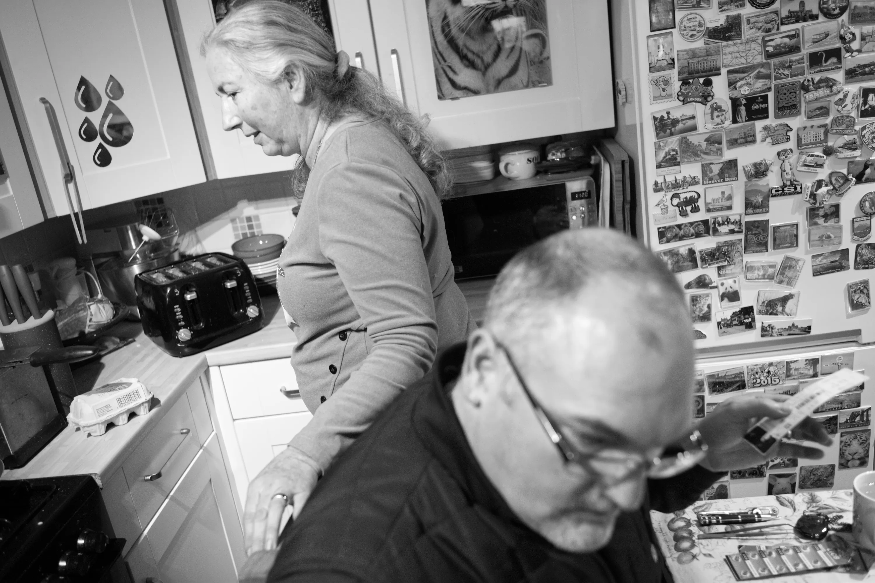 A woman standing at the kitchen counter in the foreground, a man seated in the foreground examining a small object, a fridge covered in souvenir magnets visible behind them