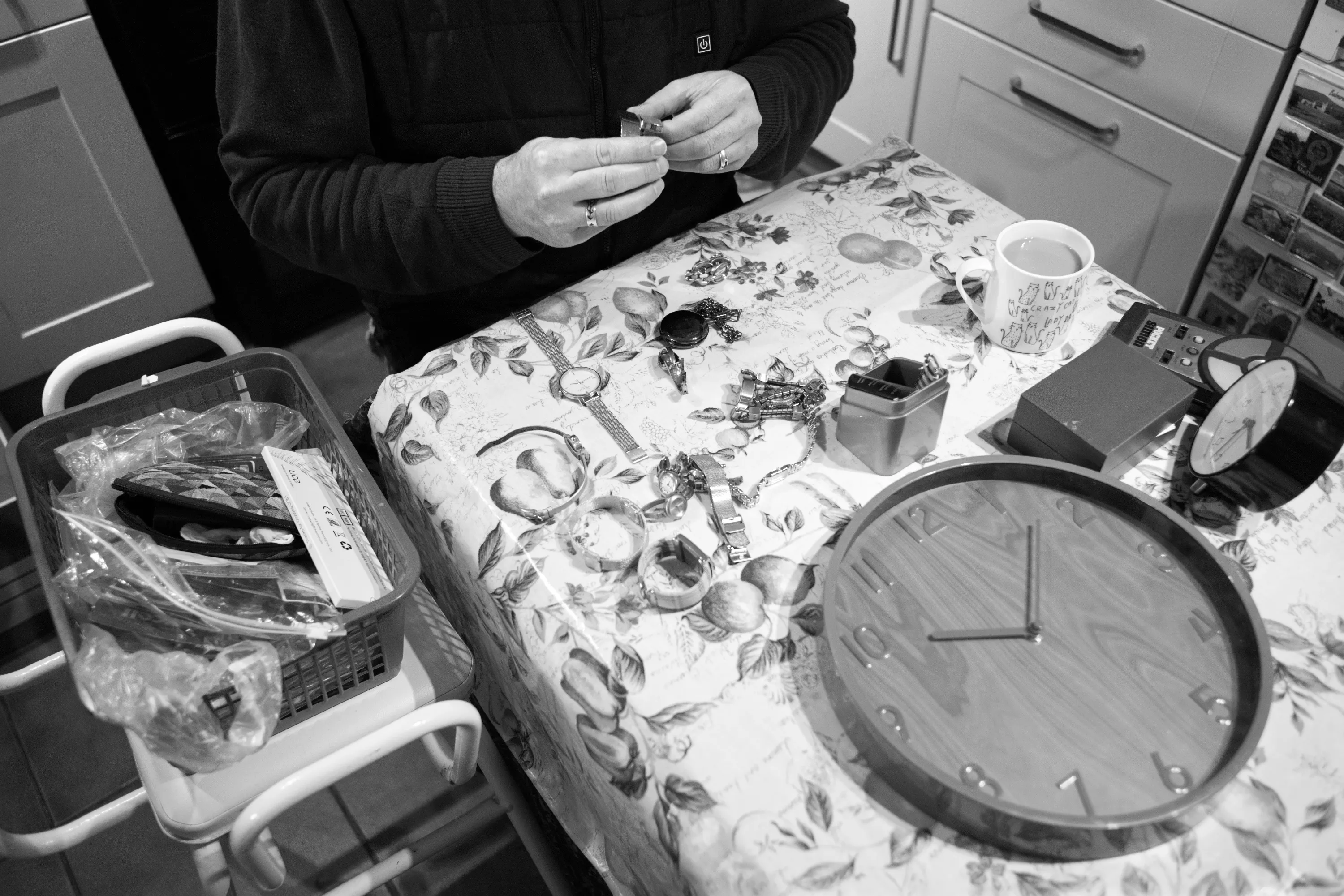 A man's hands close up, working on a small watch at the kitchen table, with several watches, a clock, tools, and a mug laid out before him