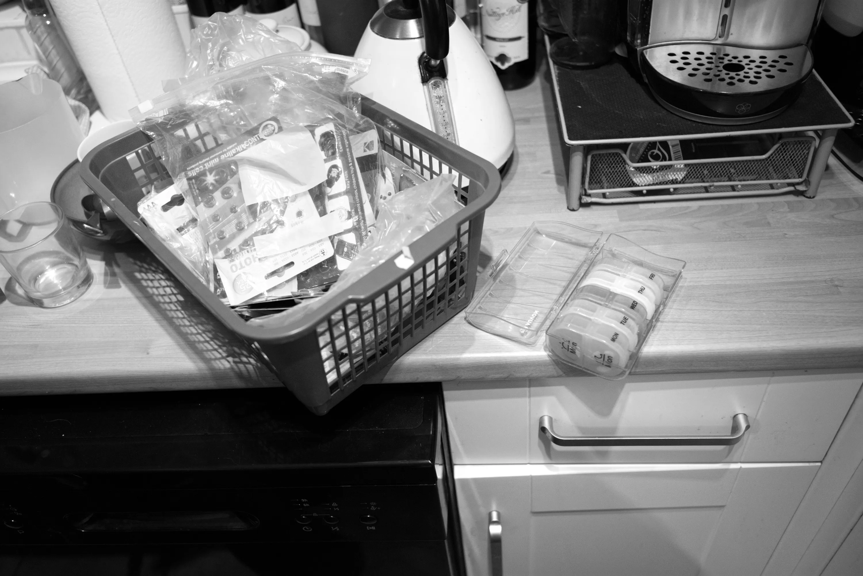 A kitchen worktop with a plastic basket filled with blister packs of medication and, beside it, a stack of transdermal patches in their packaging