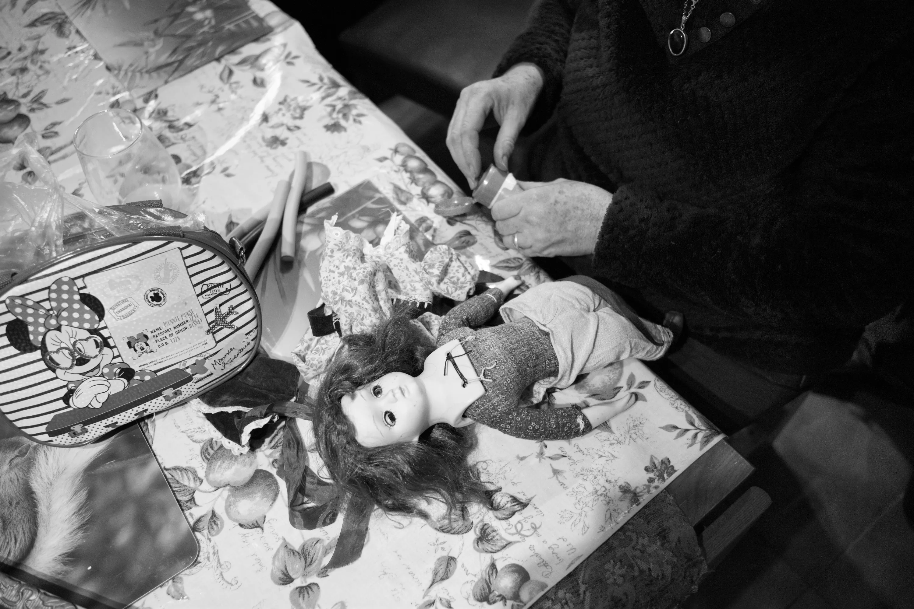 A woman's hands working on a porcelain doll lying face-up on the kitchen table, a small Minnie Mouse bag and sewing materials nearby