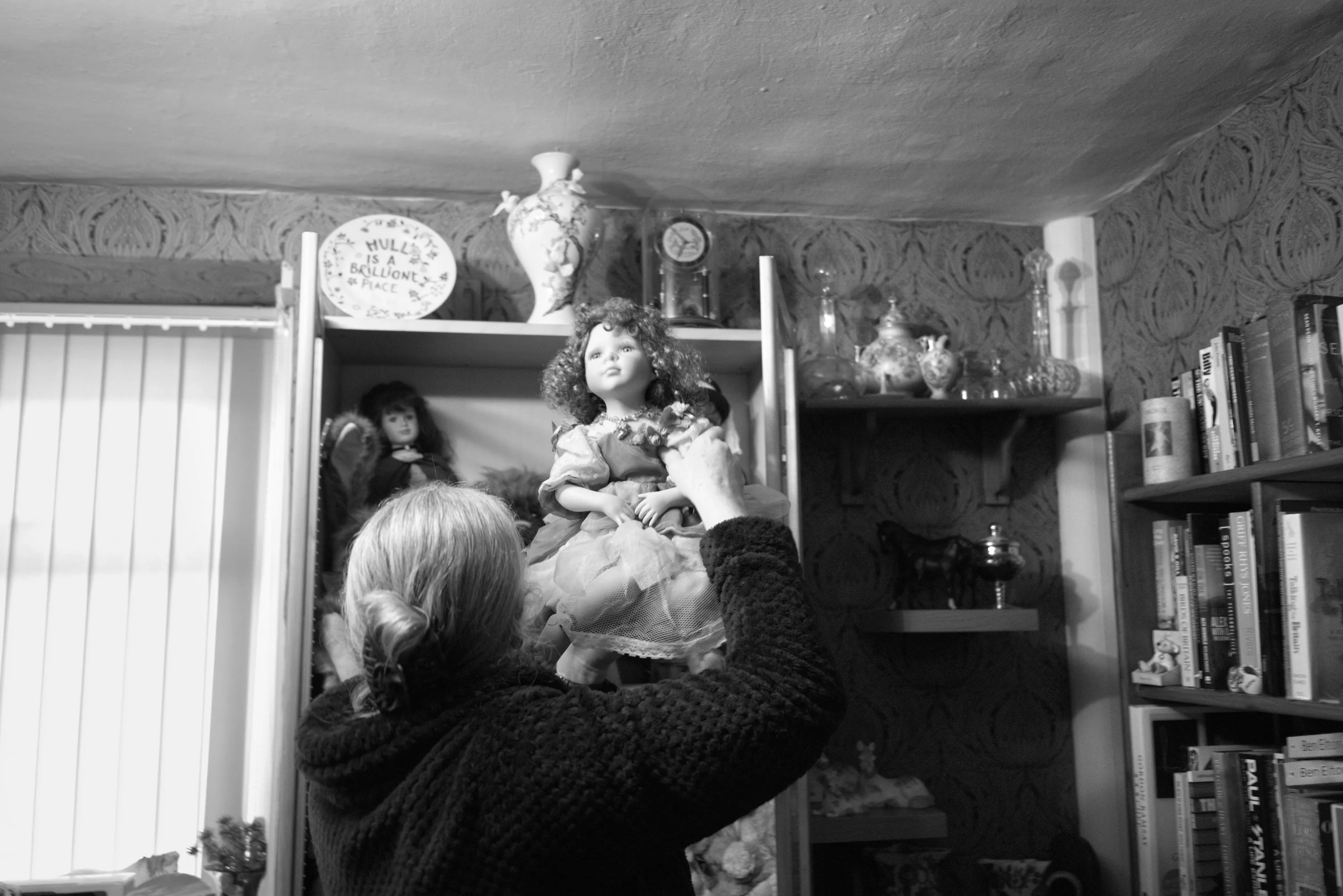 A woman lifting a large restored porcelain doll up towards a shelf in a room lined with books, ornaments, and other dolls