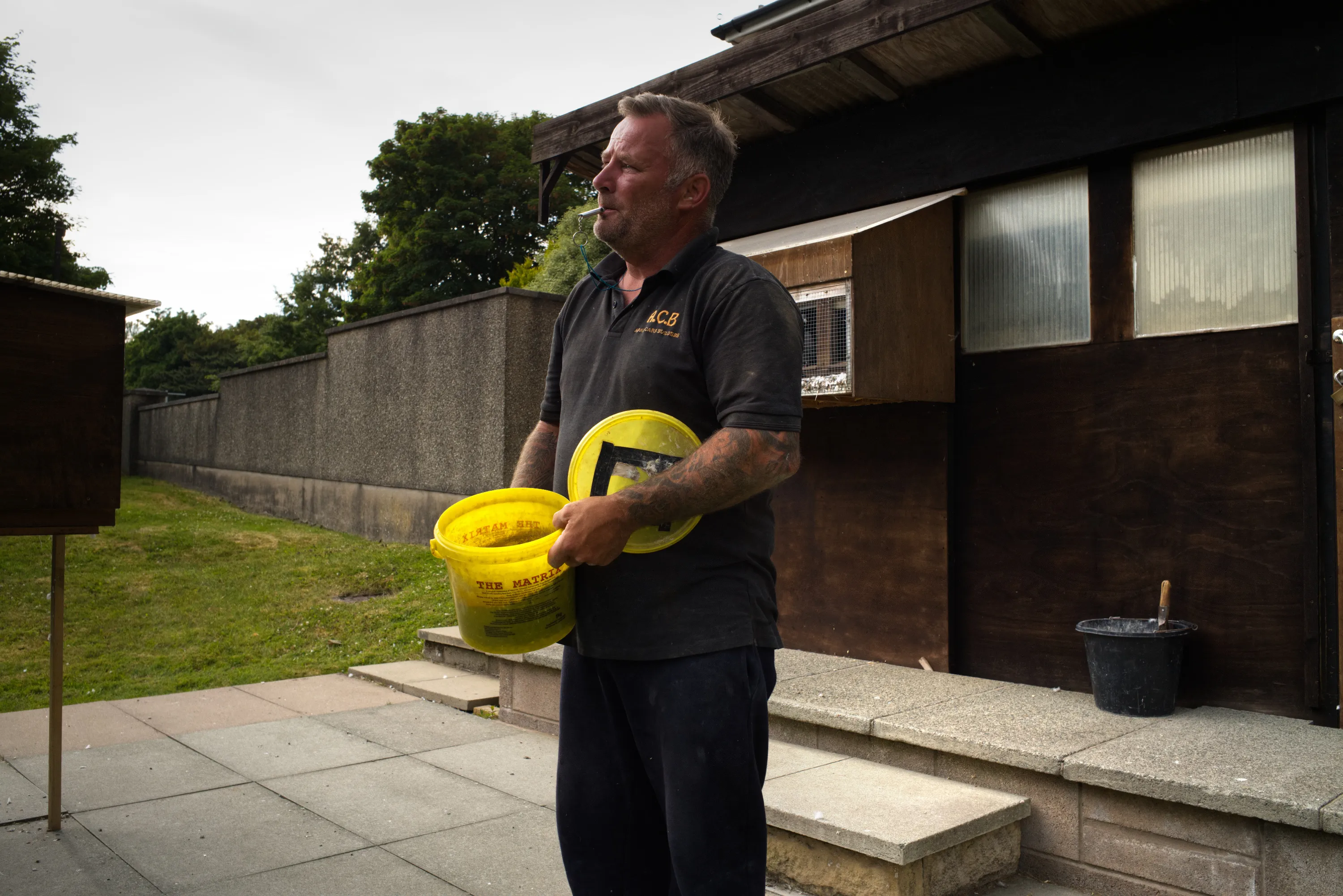 Man carrying a yellow feed bucket outside a wooden pigeon loft